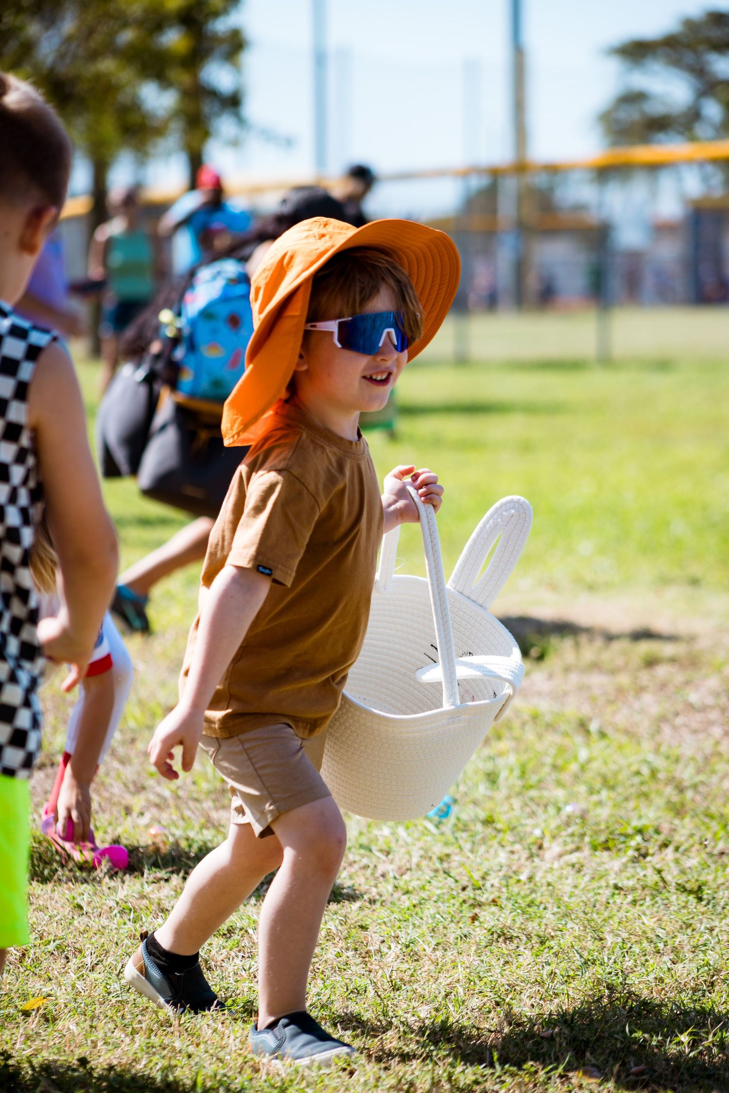 Child wearing orange sunhat and brown shirt carries white plastic Easter basket while hunting for eggs outdoors.