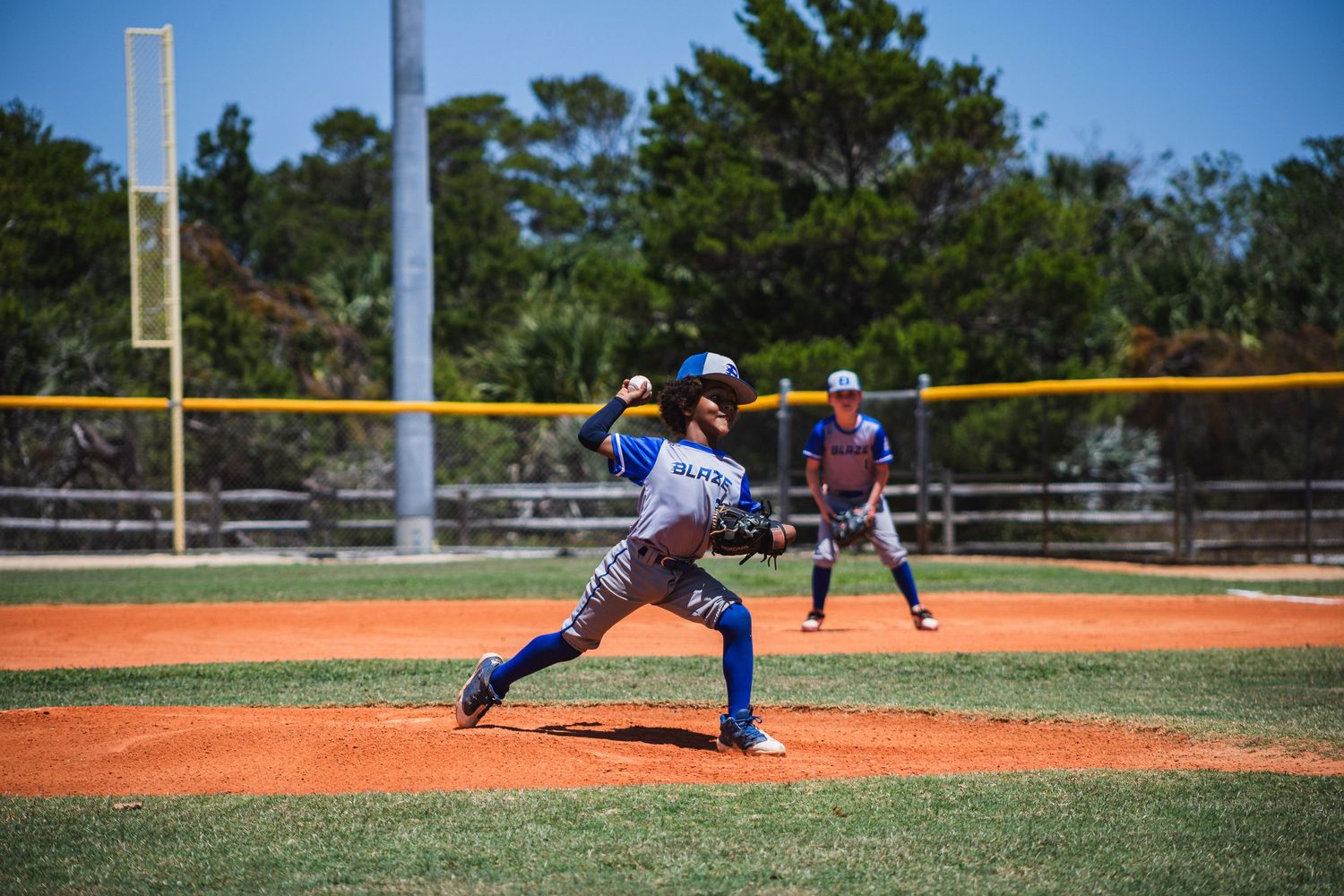 Young baseball pitcher in blue uniform throws pitch on sunny baseball field with outfielder in background.