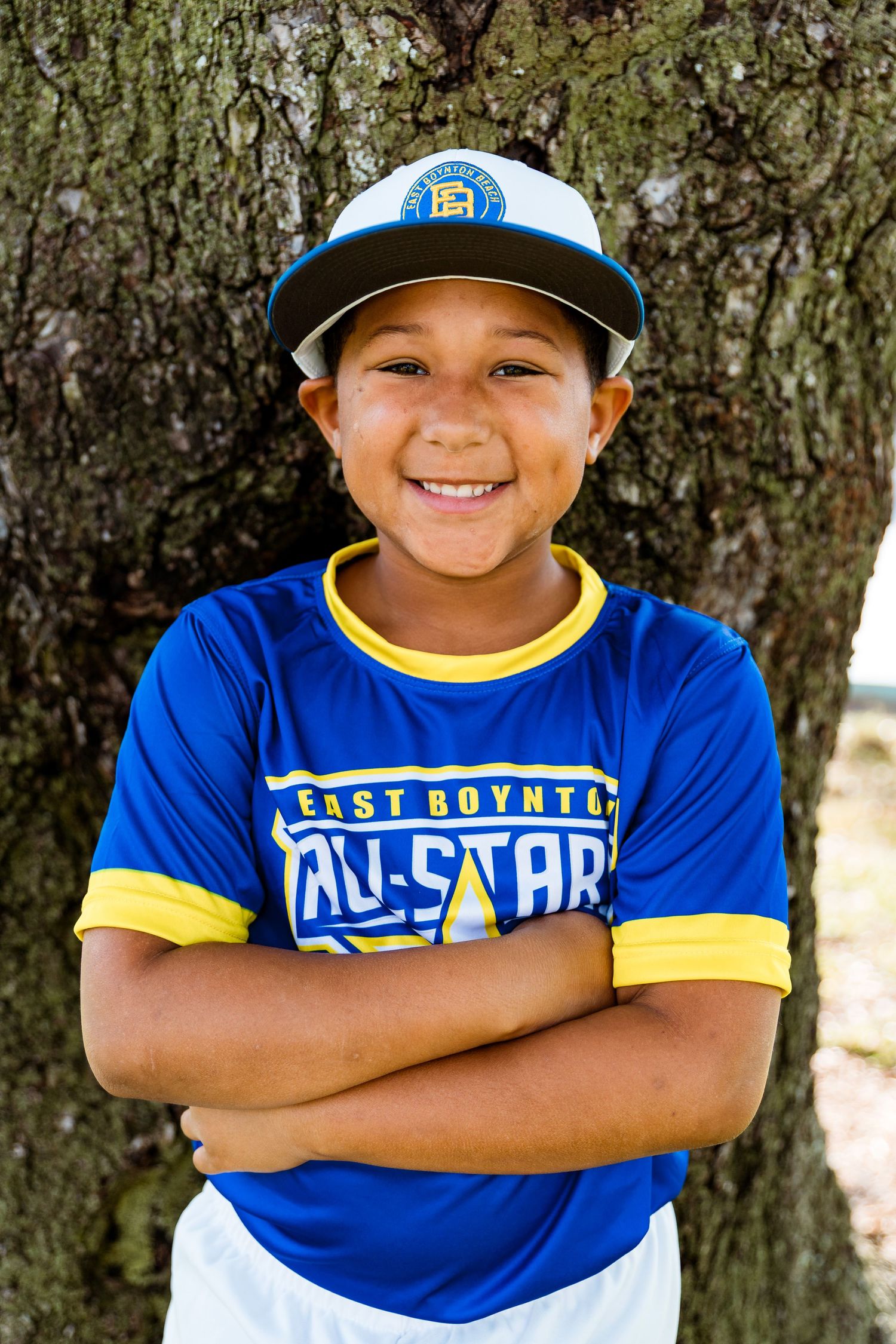 Young baseball player in blue East Boynton All-Stars uniform and cap stands with crossed arms against tree trunk.
