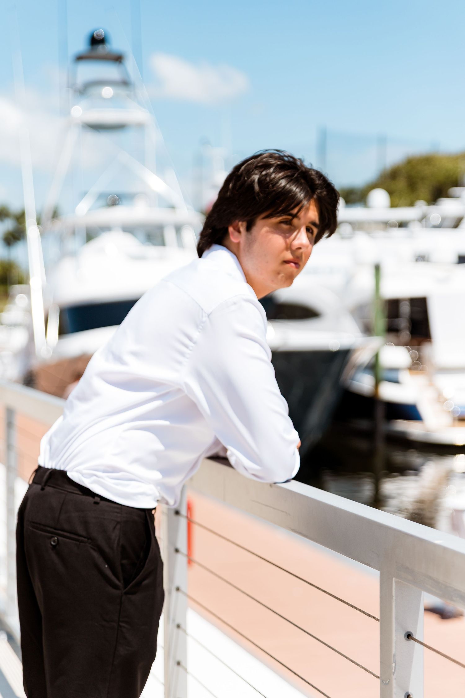Person in white dress shirt and dark pants looking over railing at marina with boats and lighthouse in background.