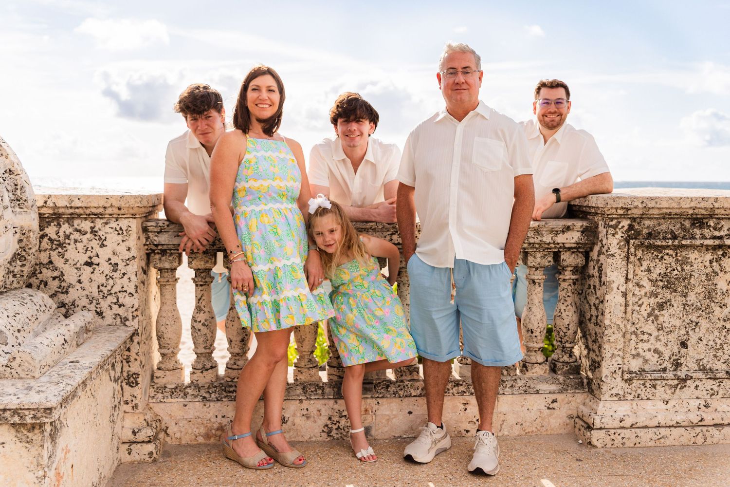 A group of people wearing coordinated outfits in blue and mint green pose together on a stone balcony overlooking the ocean.