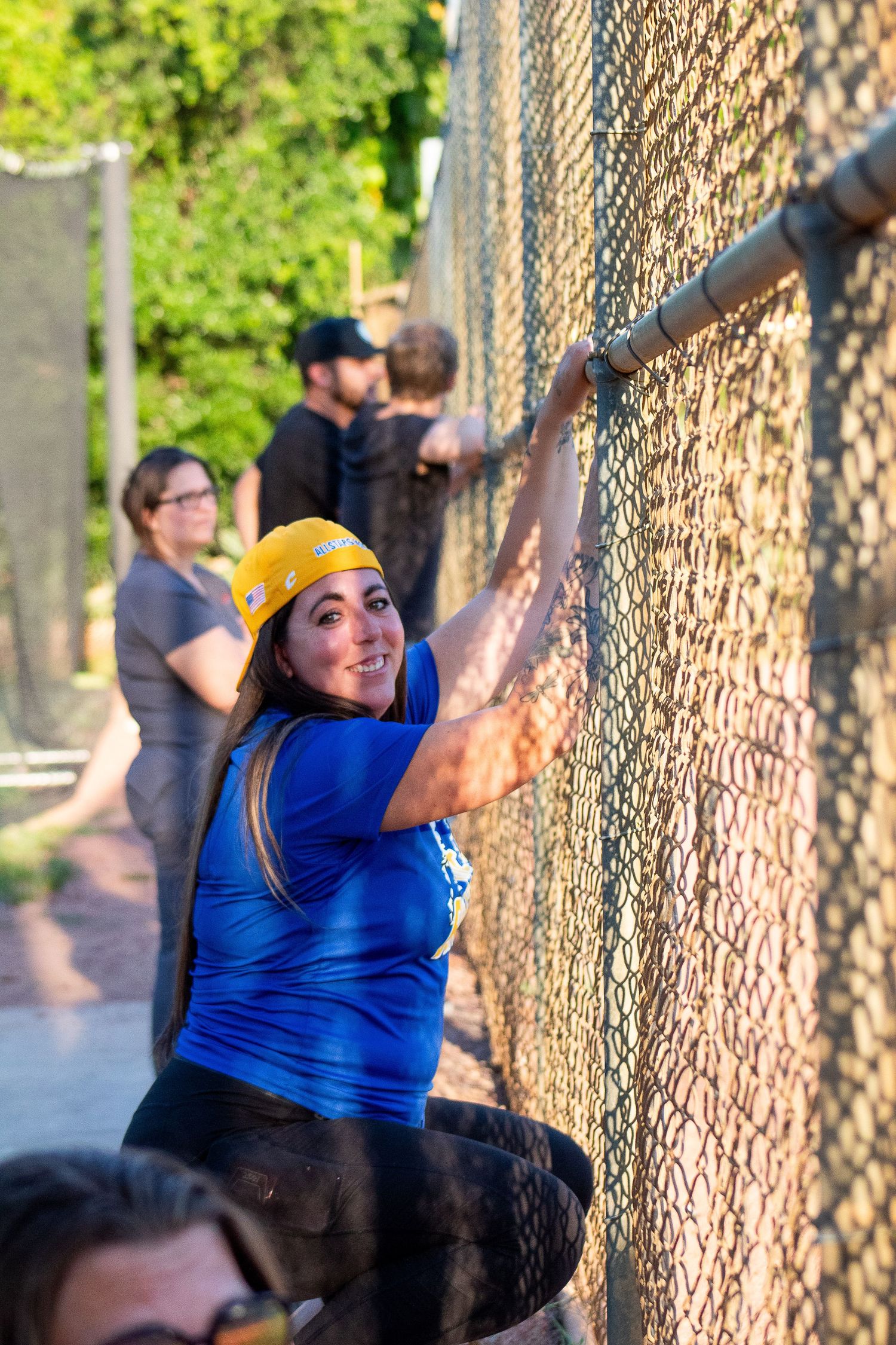 Spectator in blue shirt and yellow cap climbs chain link fence at baseball field.