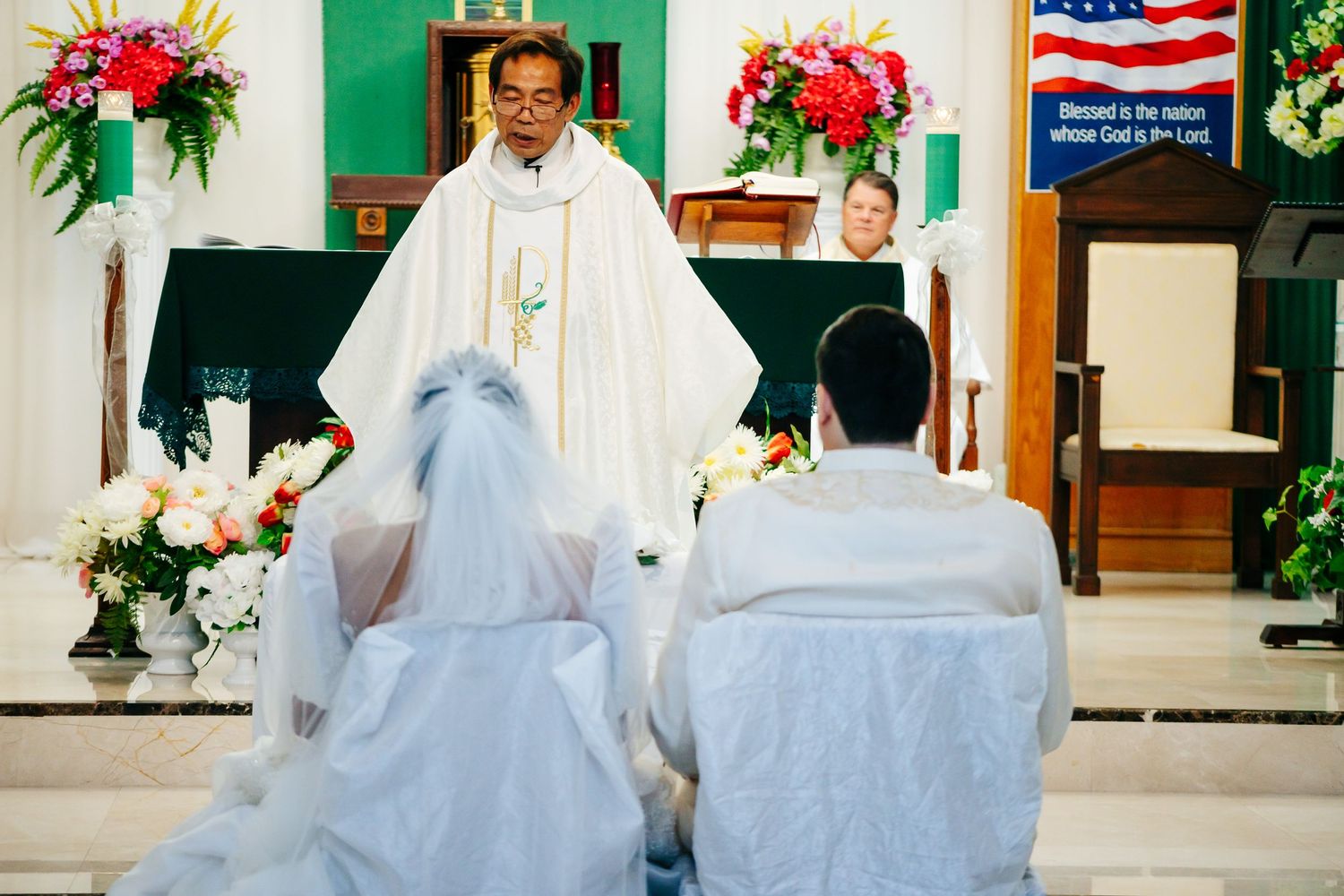 Priest performs wedding ceremony for couple kneeling at decorated church altar.