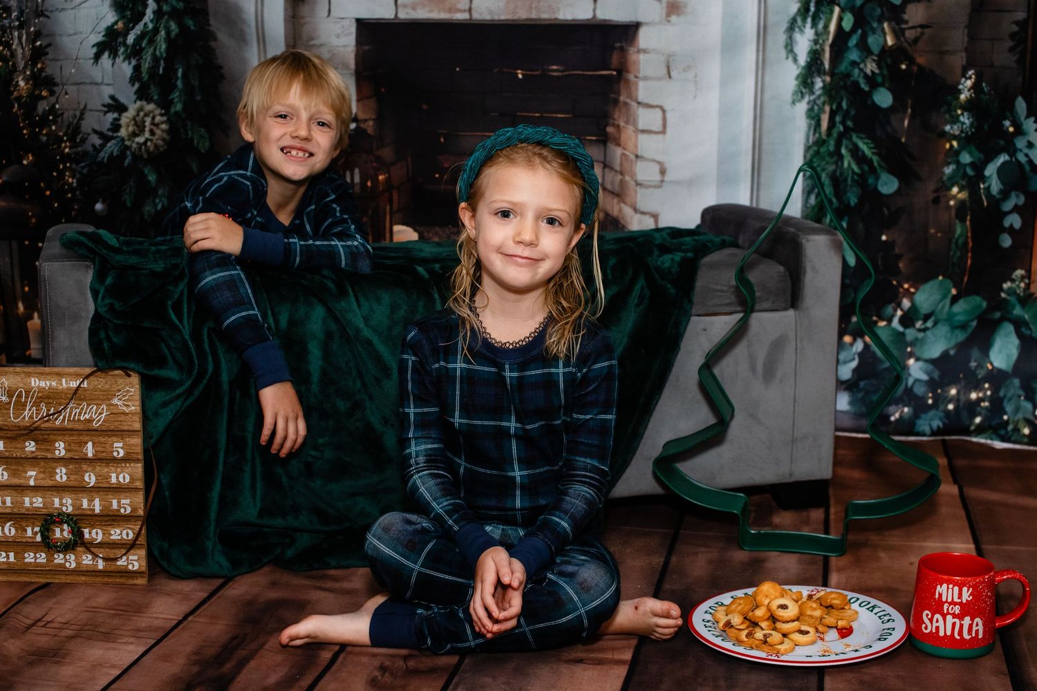 Two children in matching plaid pajamas sit on a green velvet couch by a fireplace with holiday decorations.