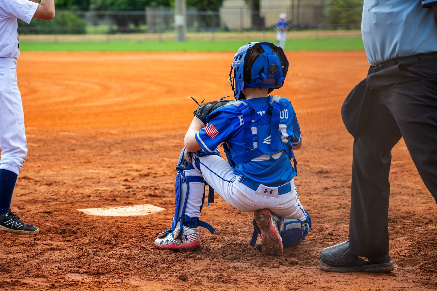Young baseball catcher in blue uniform and gear crouches behind home plate on dirt infield during game.
