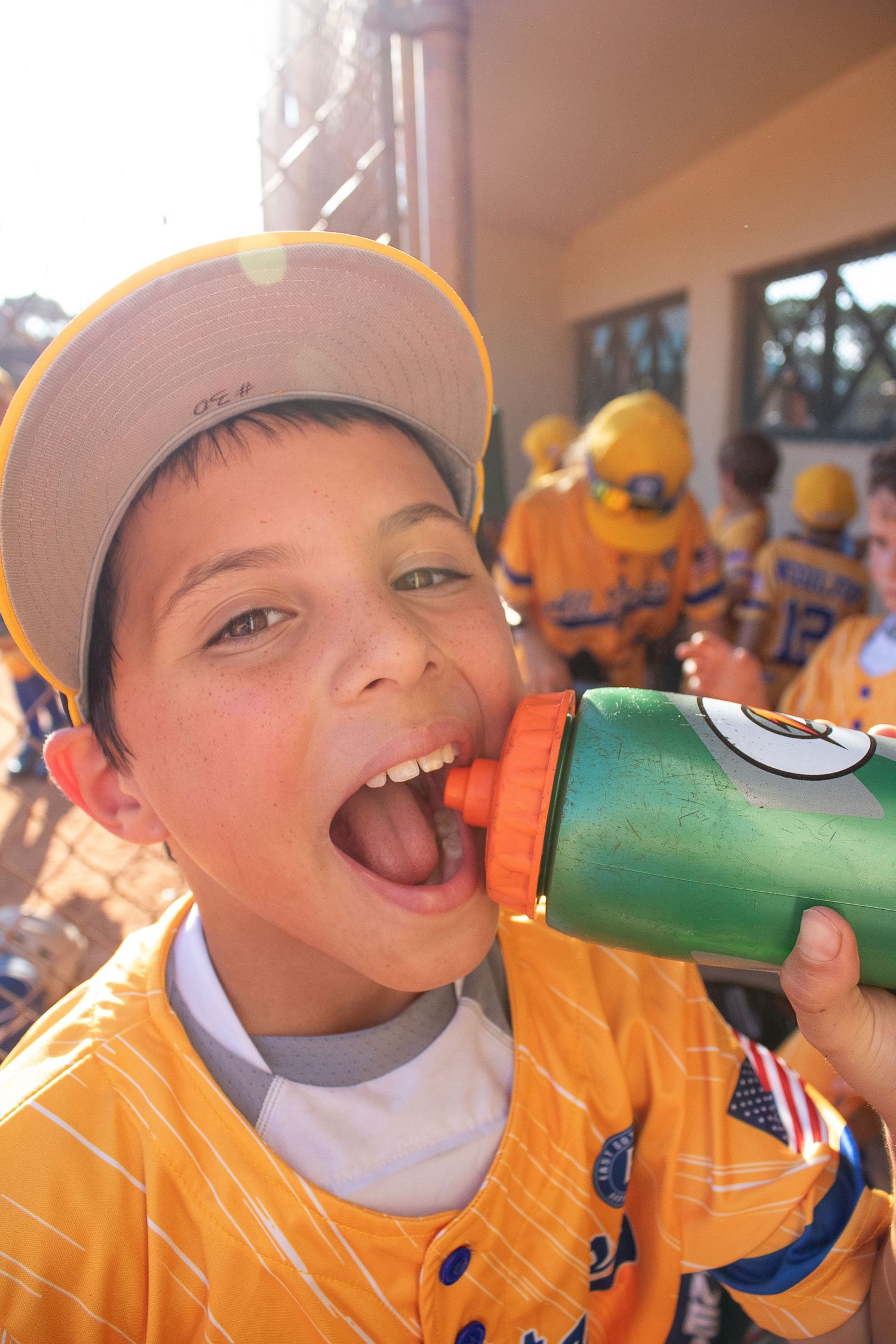 Youth baseball player in yellow uniform drinks from water bottle during game break.
