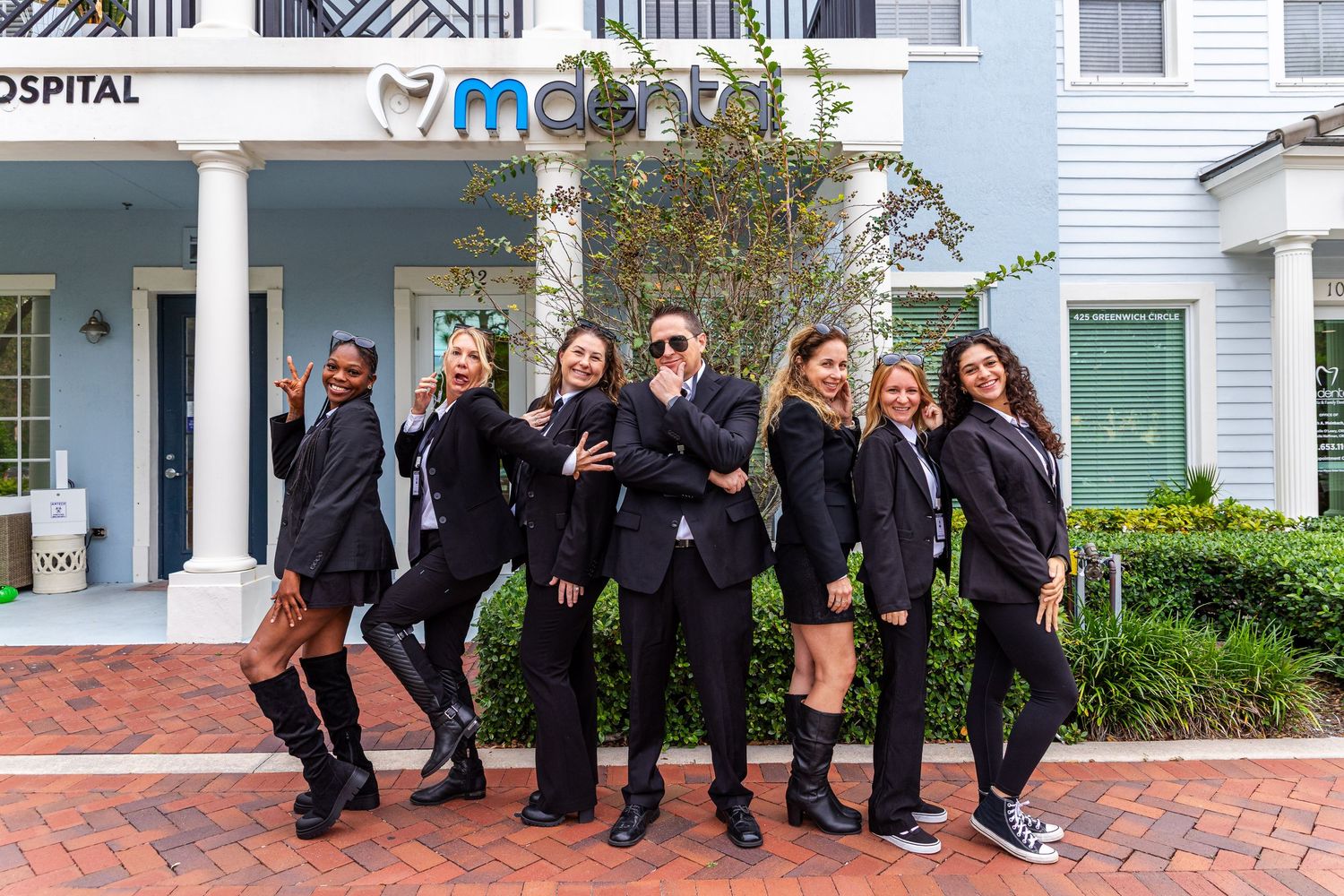 A group of people in formal attire pose together on brick steps in front of a white building.
