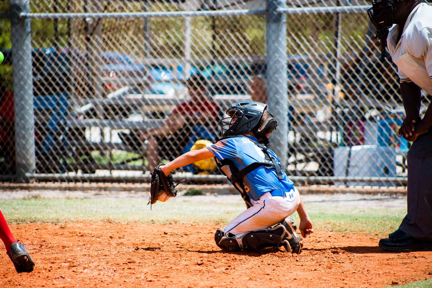 Baseball catcher lunging to catch a ball at home plate during a youth game on a dirt field.