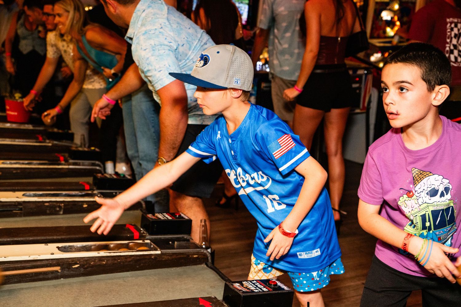 The image shows a group of young boys playing on arcade machines in what seems to be a lively, crowded arcade or gaming venue.