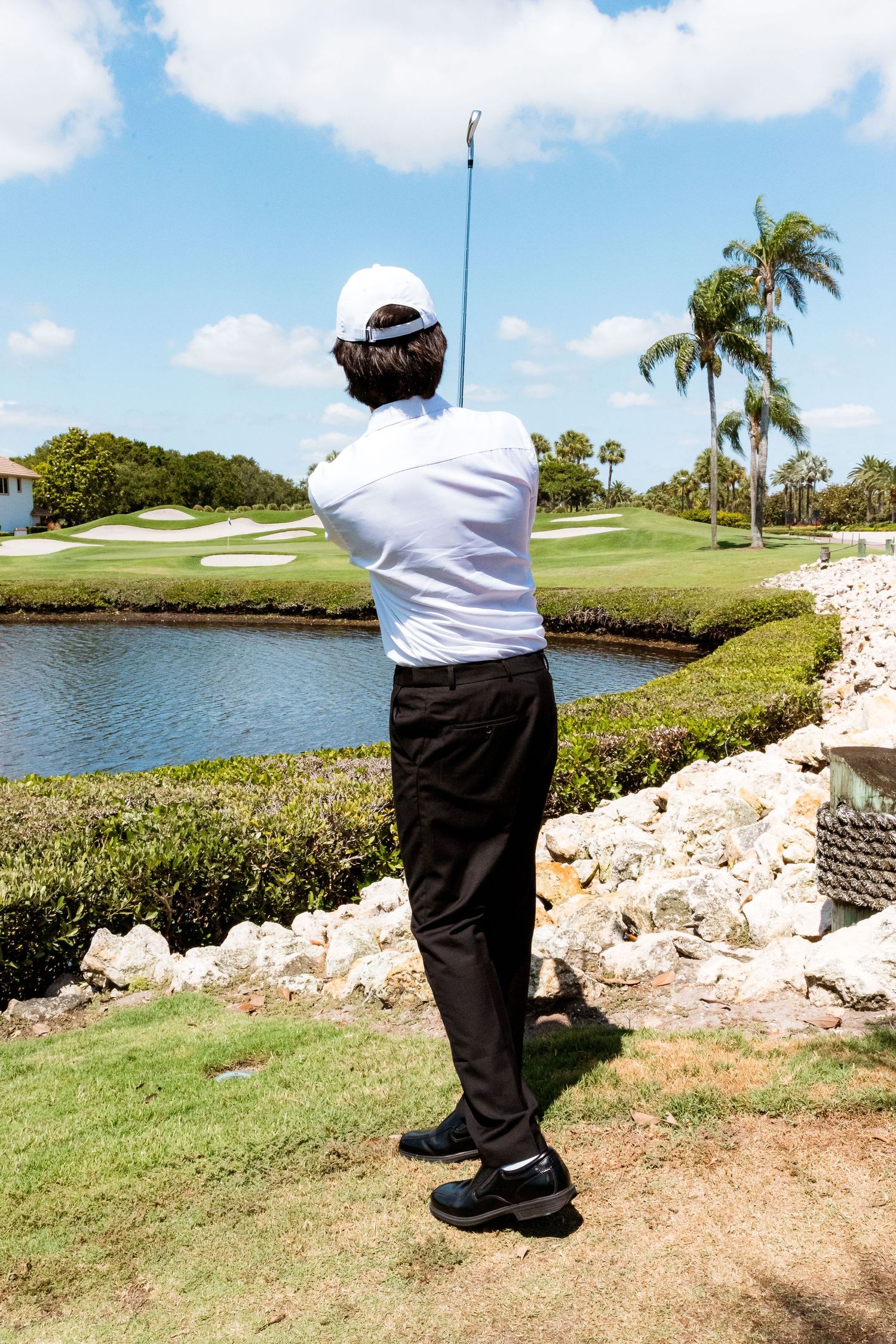 Golfer in white shirt takes a swing on a scenic course with palm trees and water hazard.