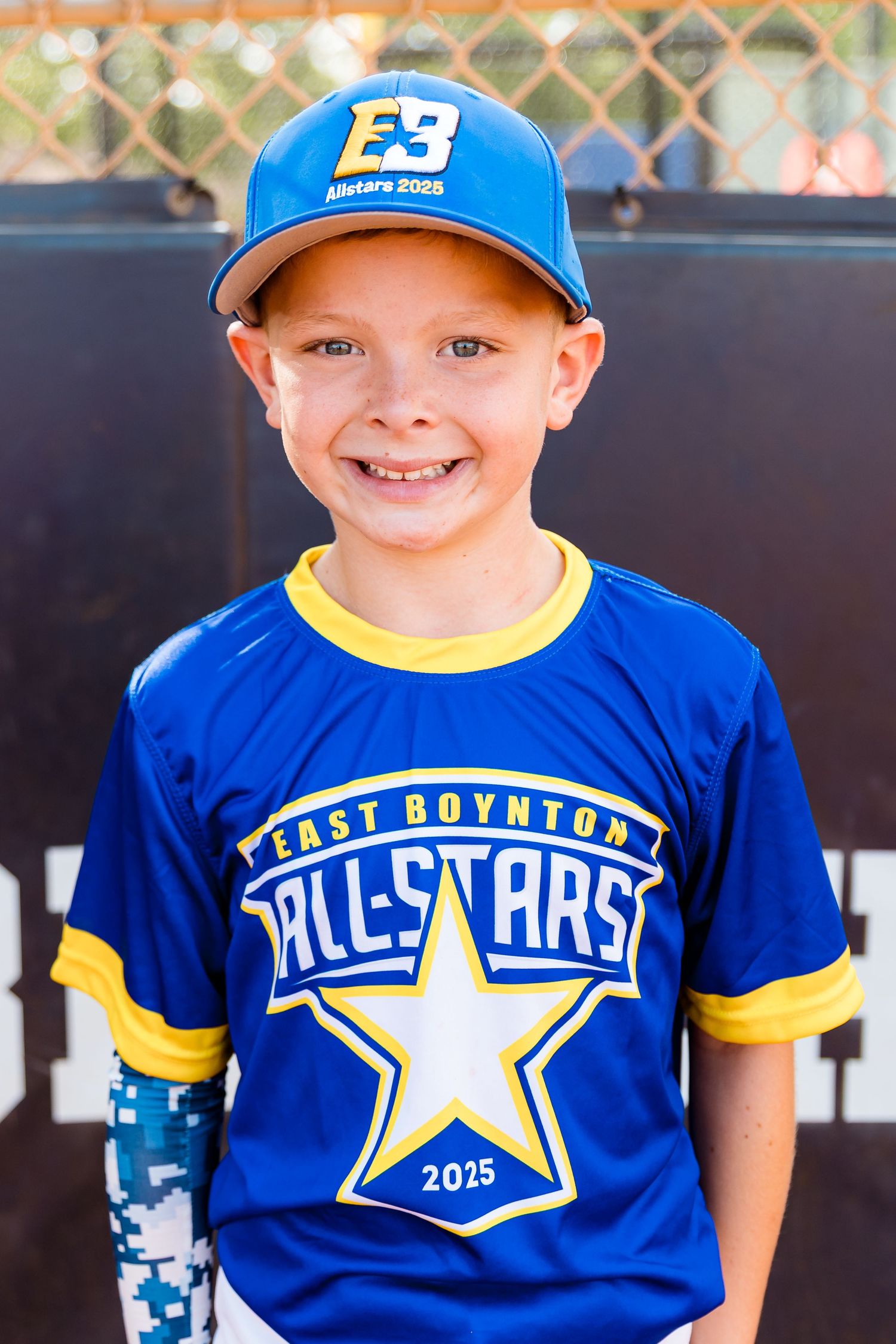 Young baseball player wearing a blue East Boynton All-Stars jersey and cap smiles brightly against a chain link fence.