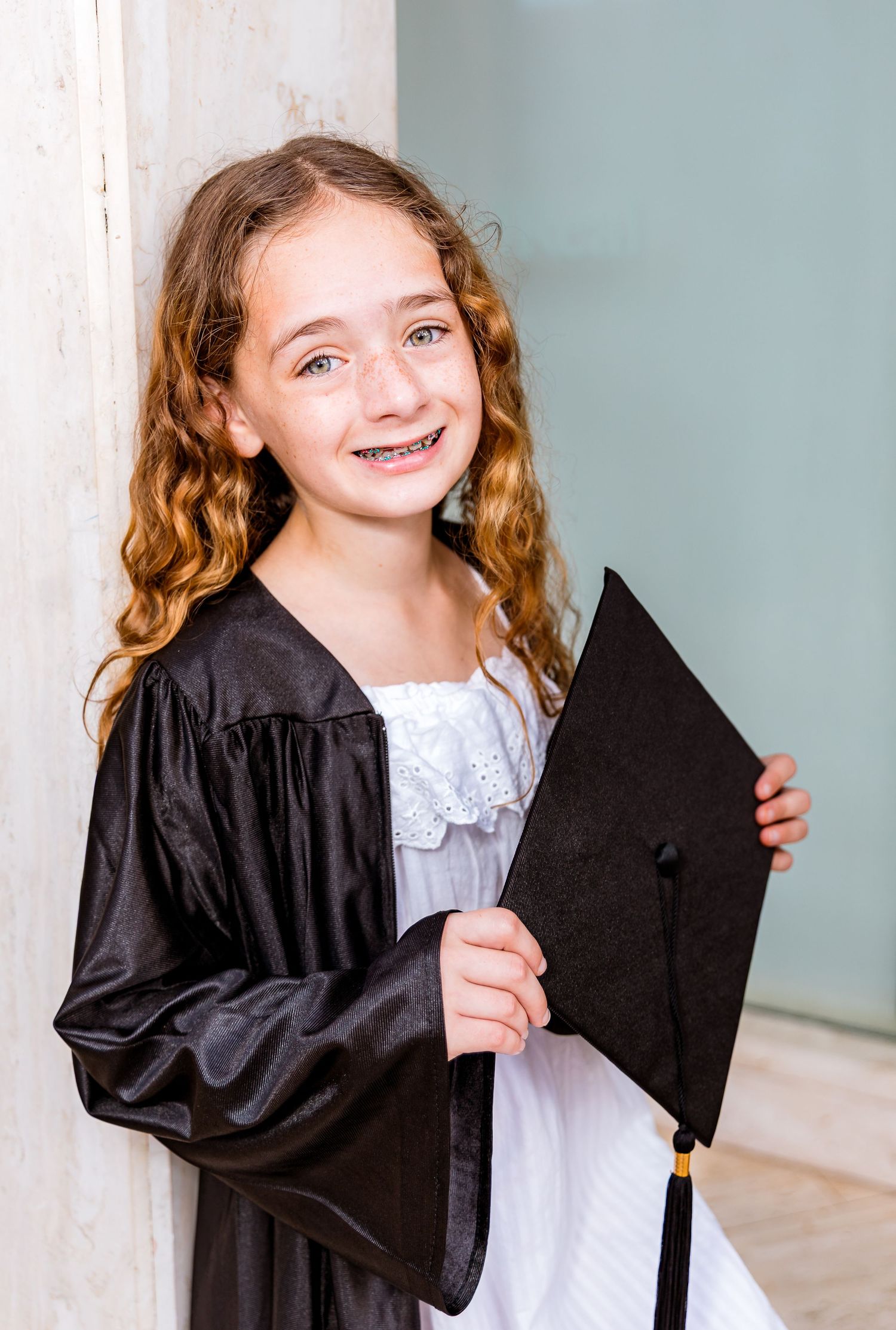 Young girl in black graduation gown and cap smiling with curly hair and braces during graduation photo.