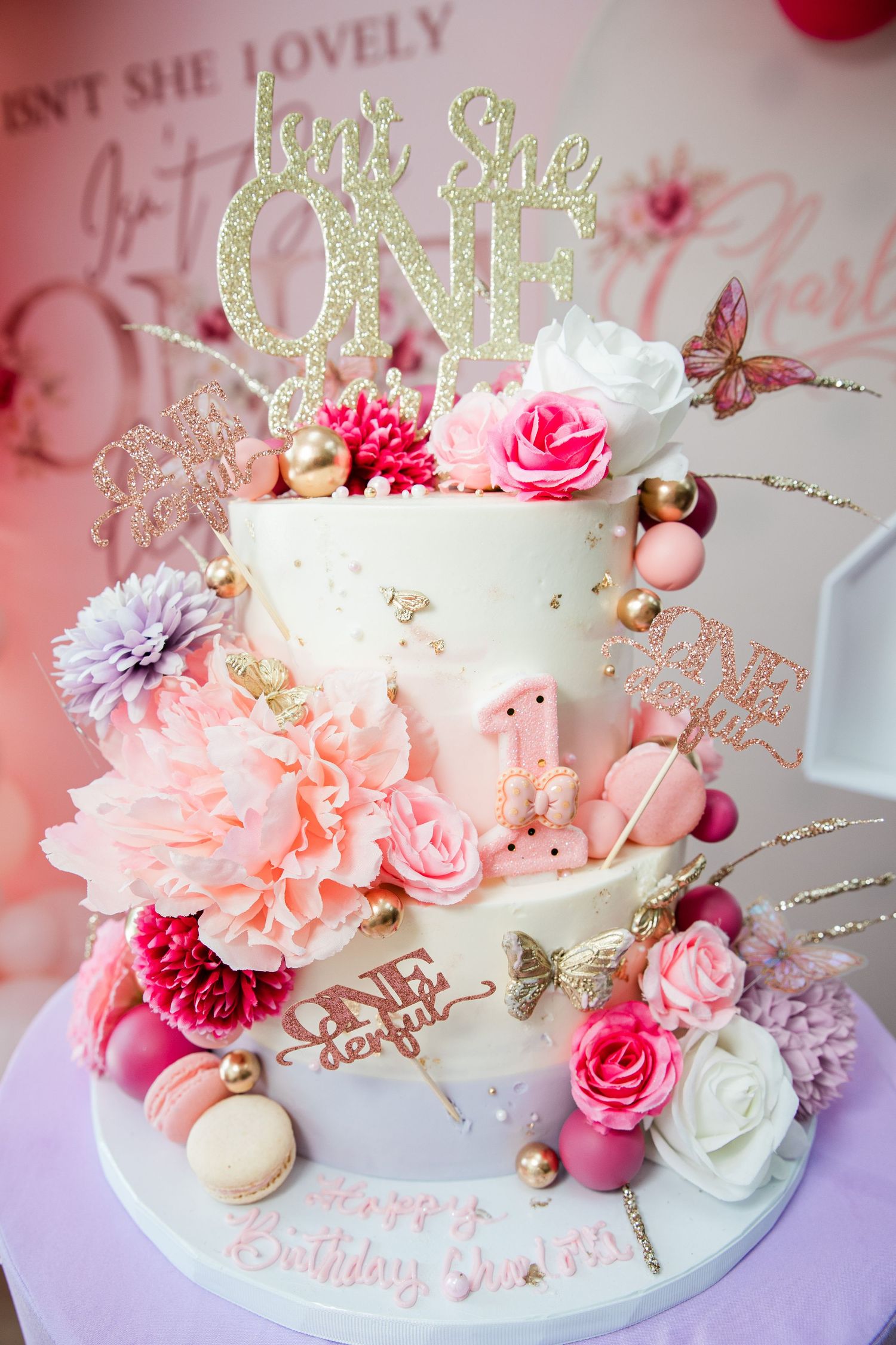 Elegant two-tier first birthday cake with pink flowers, gold accents, macarons, and “Isn’t She ONE” topper.