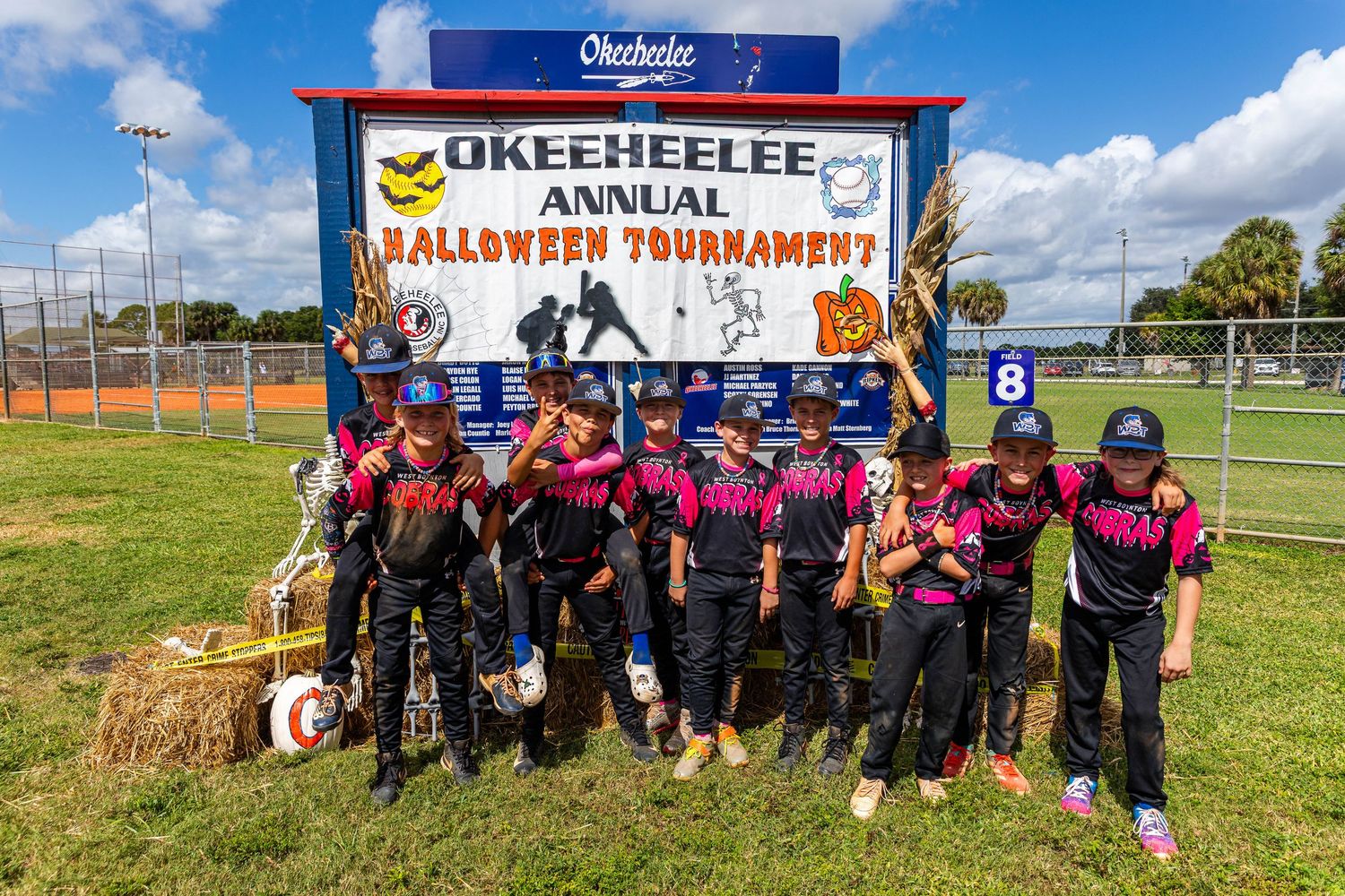 Youth football players and coaches gather on a sunny field for a team photo during practice or game day.