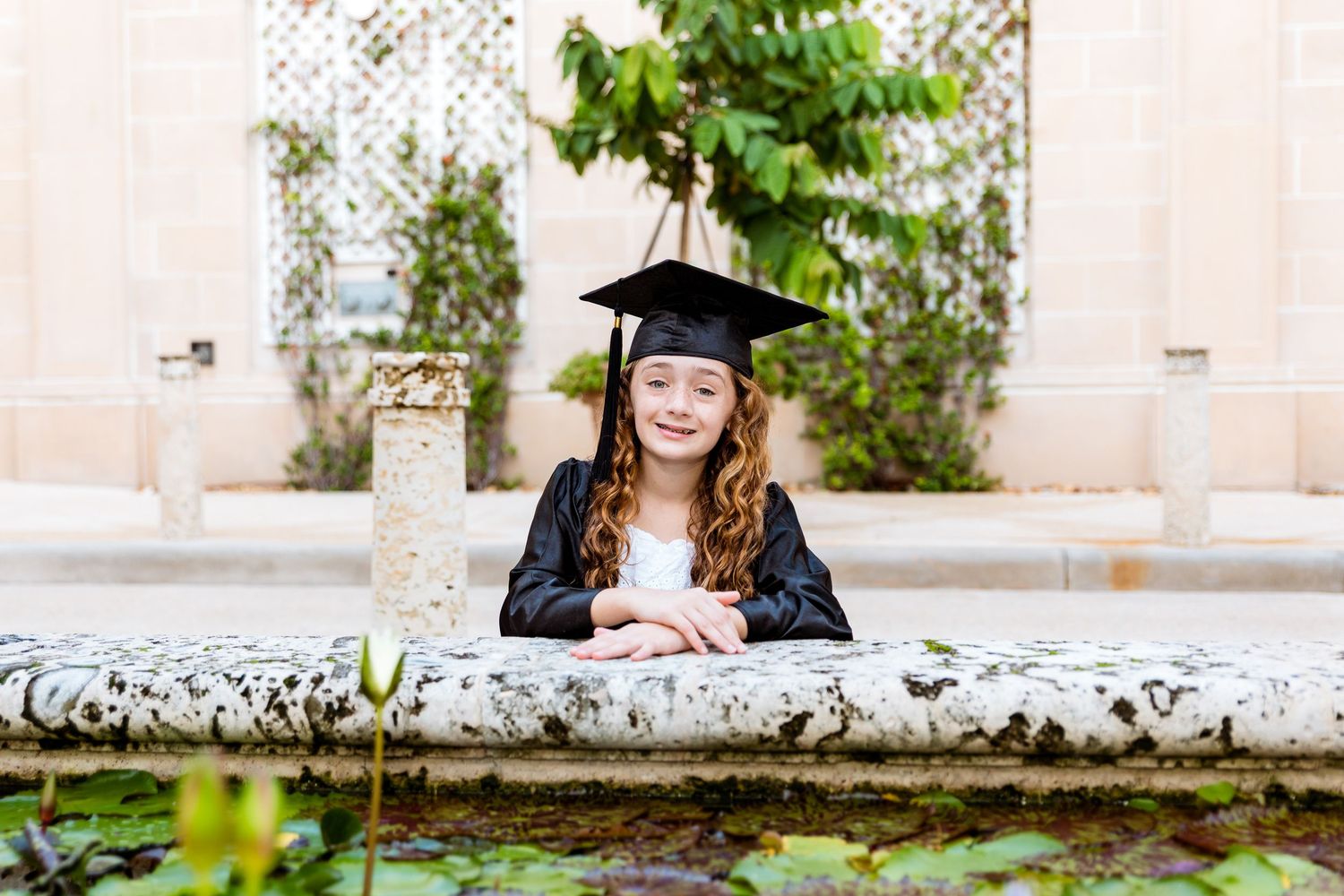 Young girl in black graduation cap and gown posing proudly by a stone fountain in a garden setting.