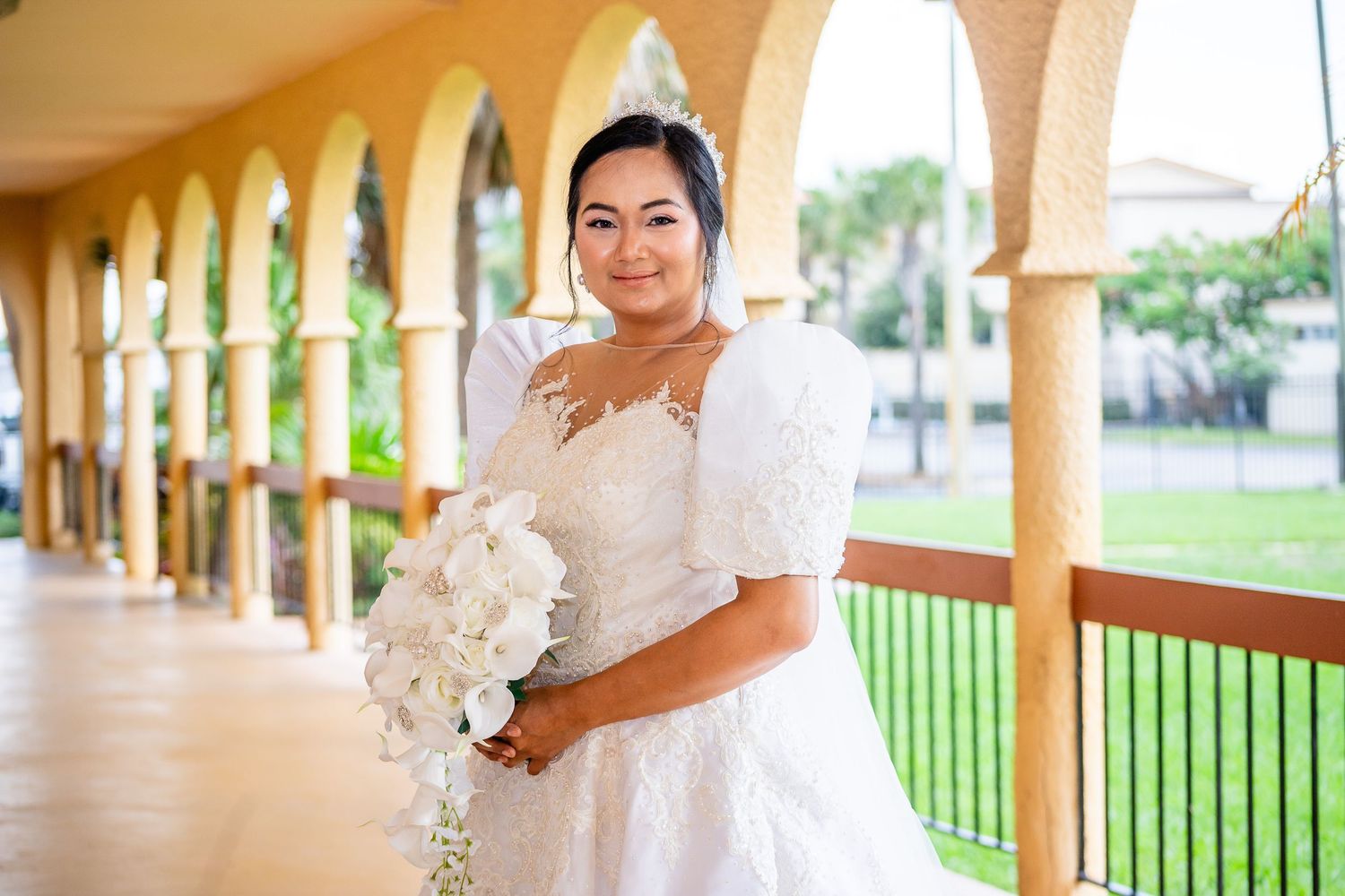 A bride in a flowy white wedding gown poses in an outdoor covered walkway with stone columns.