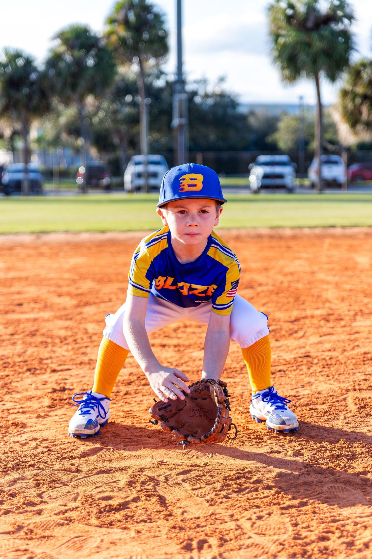A young baseball player in blue and yellow uniform practices fielding ground balls on an orange clay infield.