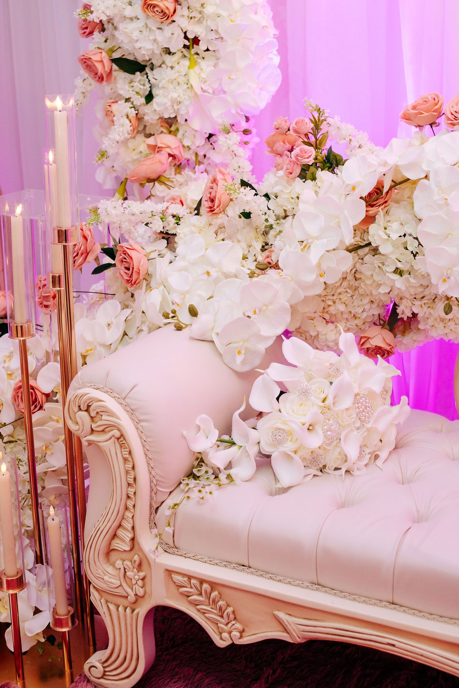 Ornate cream chaise lounge decorated with lush white and pink floral arrangements and gold candelabras against pink lighting.