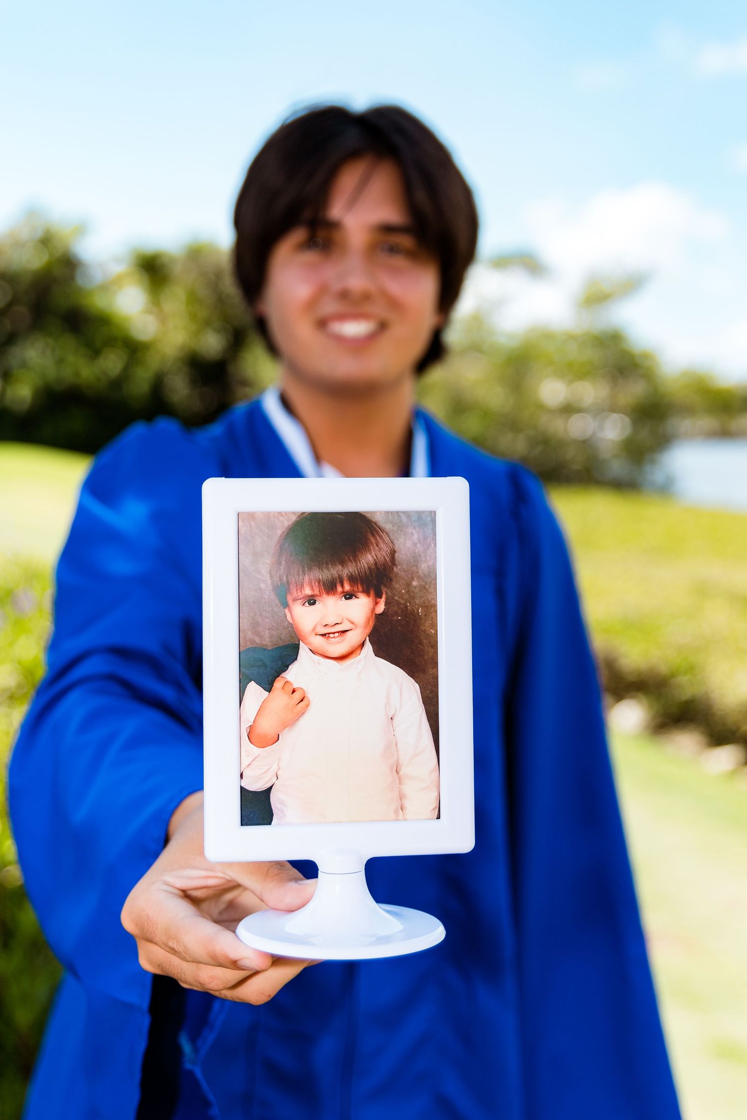 Person in blue graduation gown holds up childhood photo displayed in white frame outdoors on sunny day.