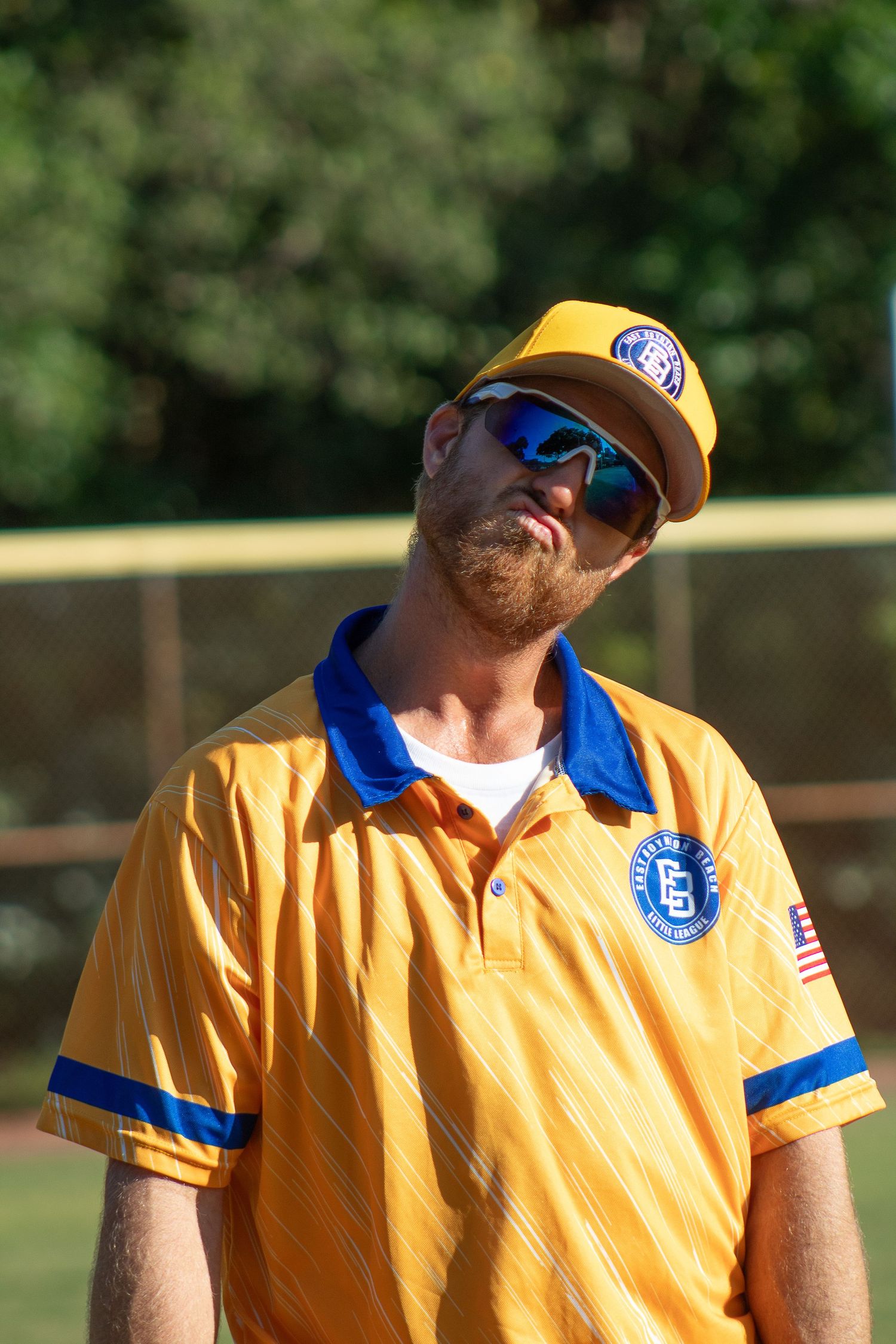 Coach wearing yellow and blue uniform with team logo and cap stands on baseball field.