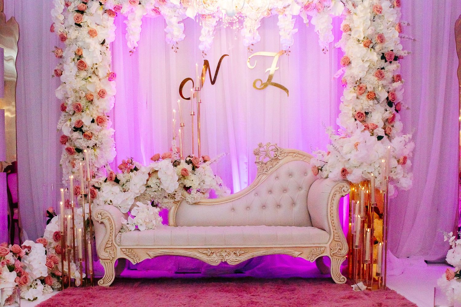 Romantic wedding photo area with white chaise lounge and floral arch against purple backdrop.
