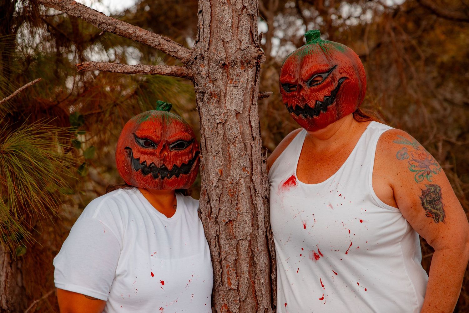 Two people wearing pumpkin masks and white shirts with fake blood splatter pose against a tree.