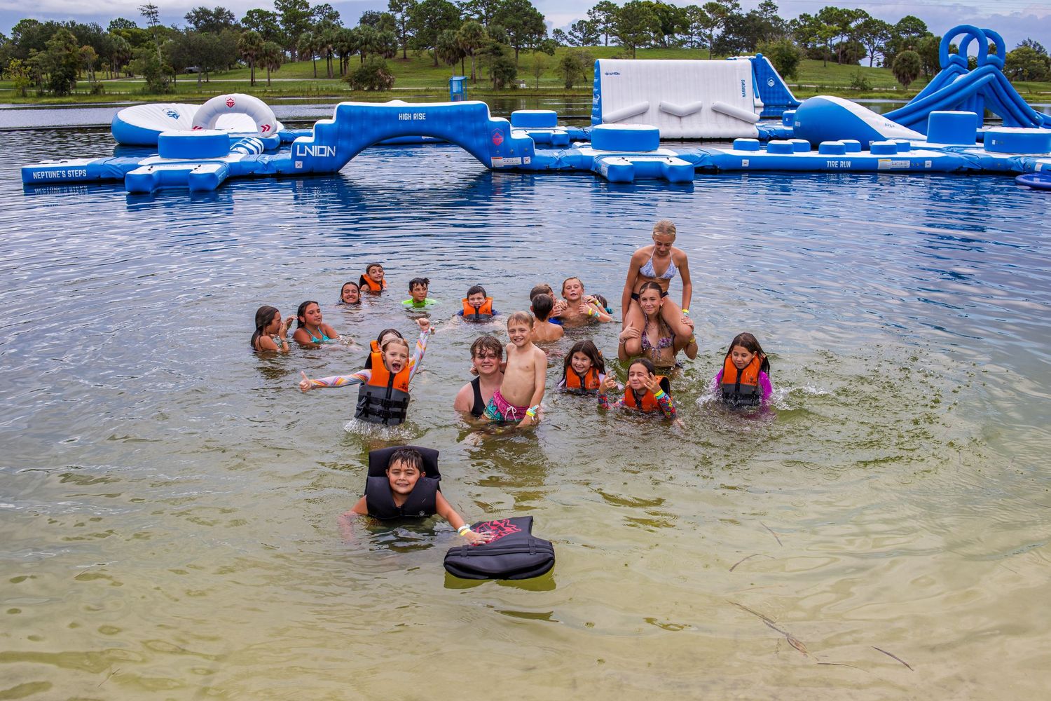 Large group enjoying recreational activities in shallow lake water with inflatable floats visible.