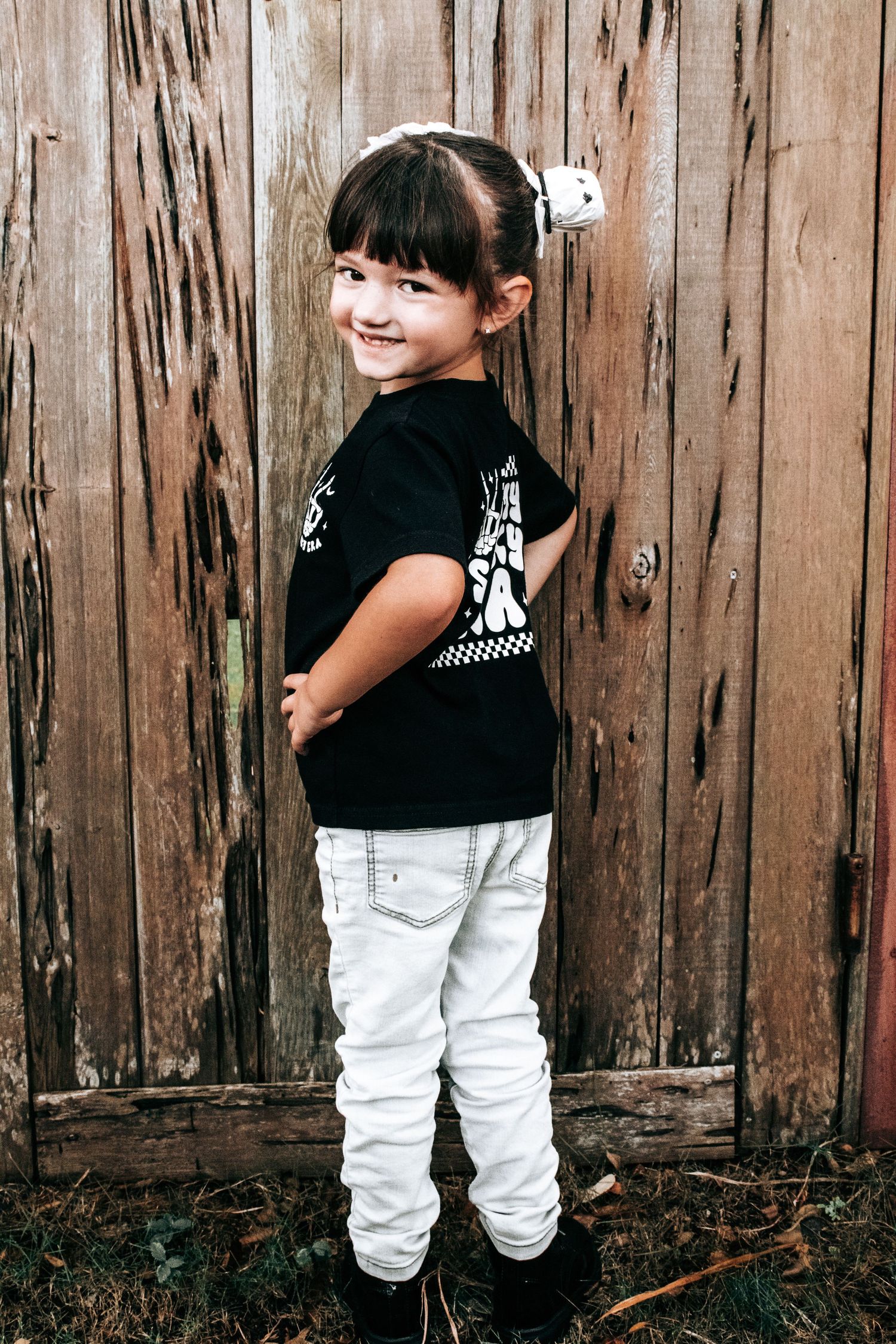 Young child in black t-shirt and white pants posing playfully against a rustic wooden fence.
