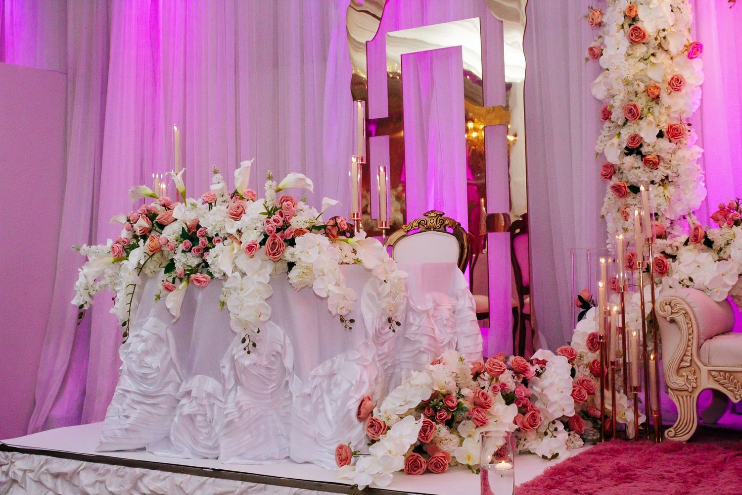 Elaborate white and pink floral arrangements with gold accents on tiered wedding stage display.