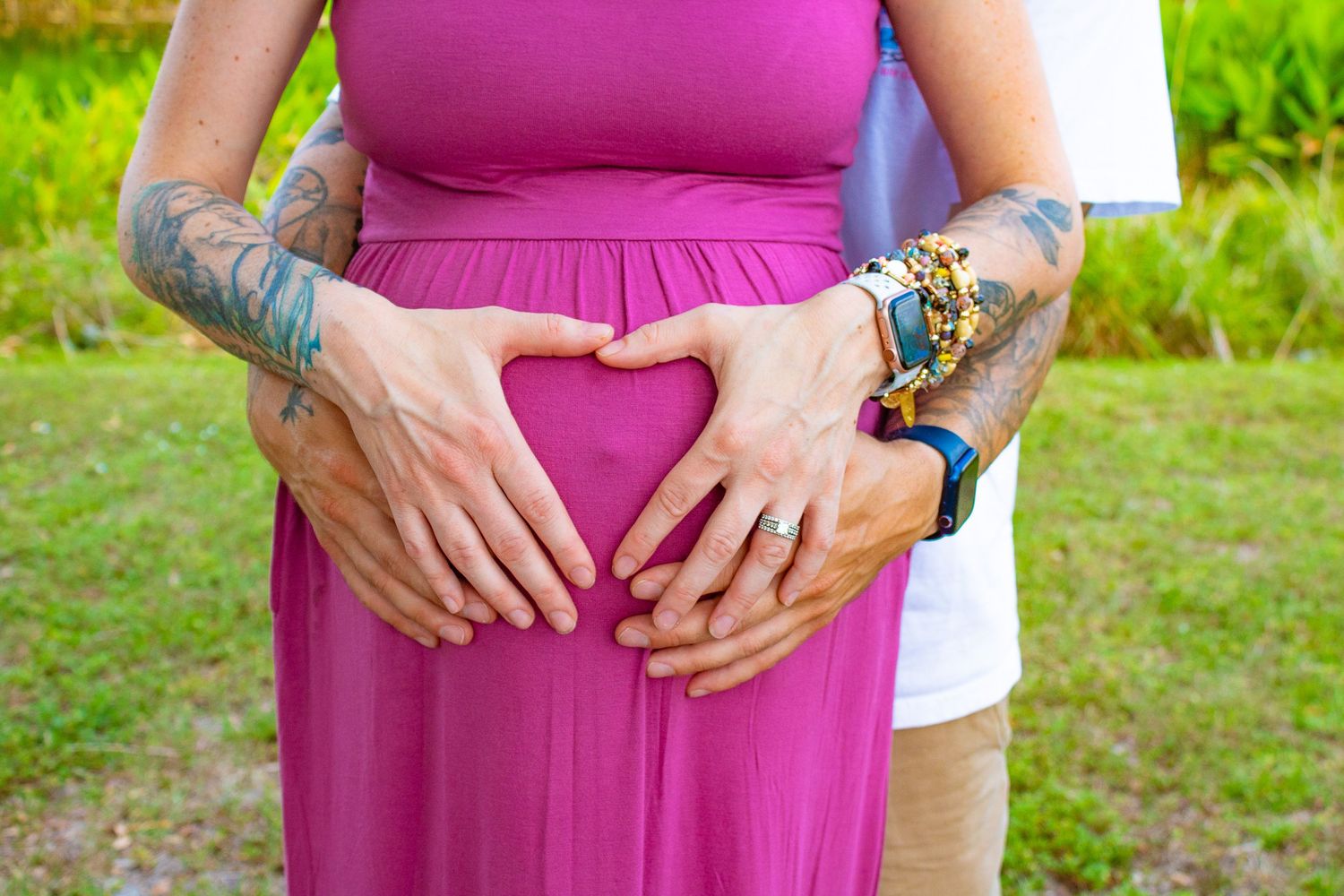 Tattooed hands creating heart shape over magenta maternity dress outdoors.