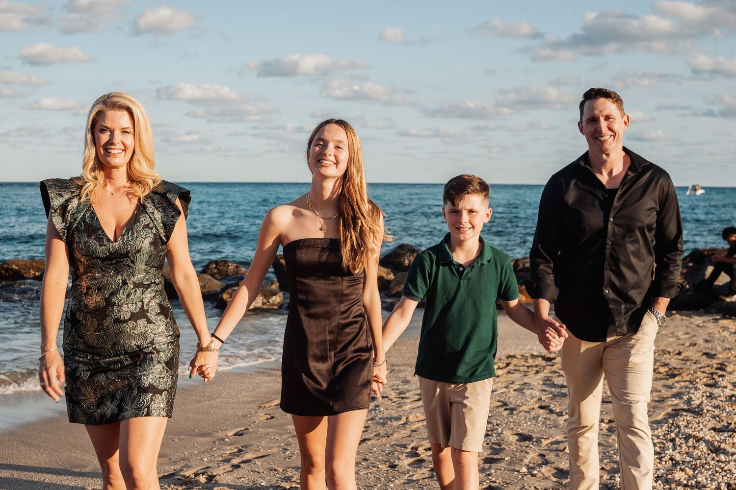 Family posing together on sandy beach with ocean waves and rocky shoreline in background.