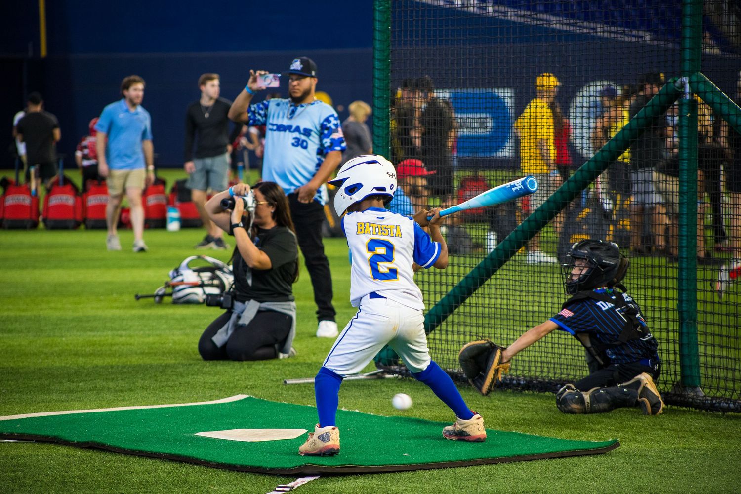 Youth baseball player takes batting practice in cage while photographers capture the moment.