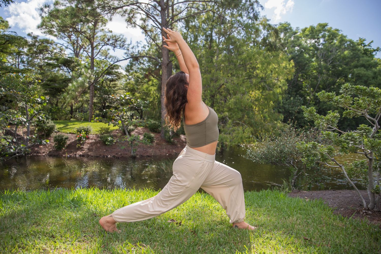 Someone performs a yoga warrior pose on green grass near a peaceful pond in a garden setting.
