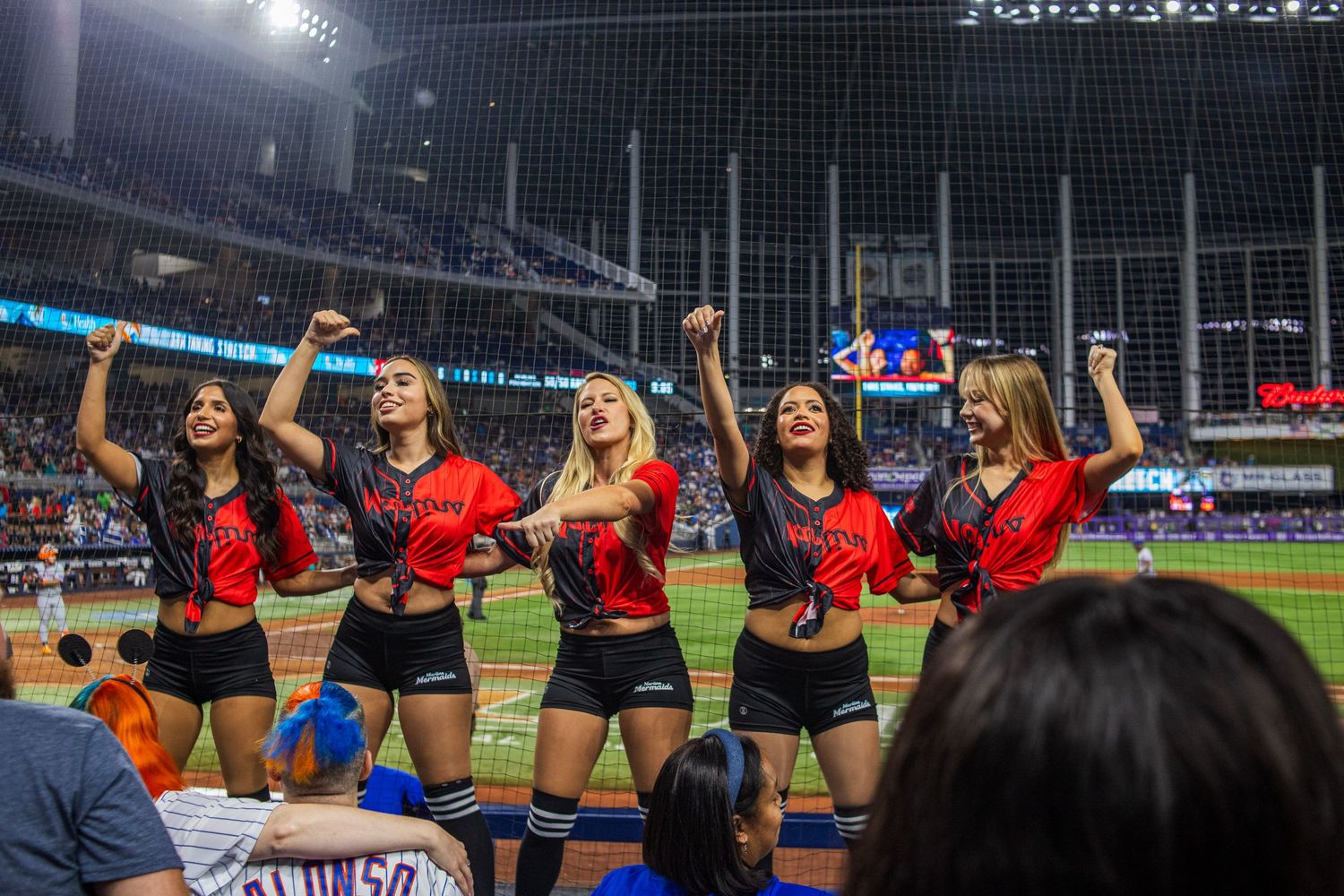 Cheerleaders perform on the field during a baseball game with stadium lights shining in the background.