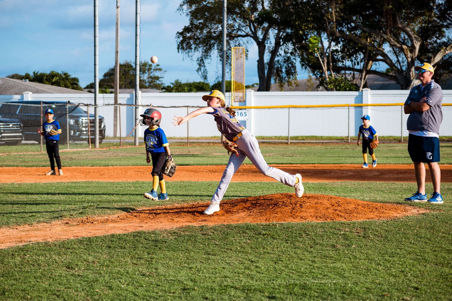 Baseball pitcher in mid-delivery on mound with coaches and players watching in background.