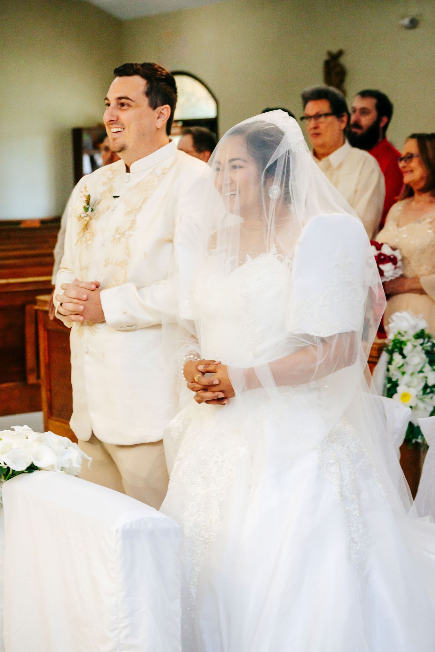 A couple stands at the altar during their wedding ceremony, with the bride in white dress and veil and groom in white suit.