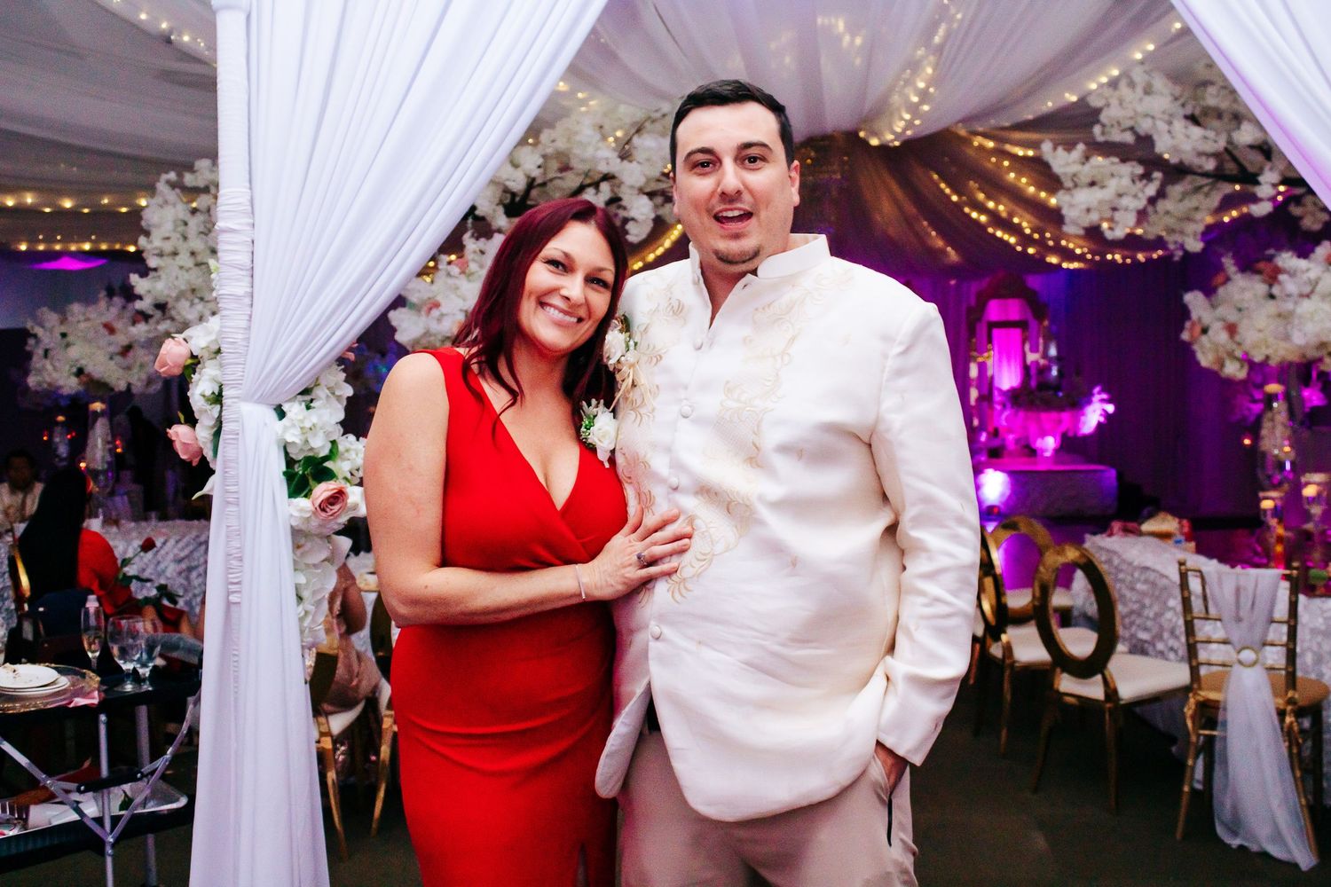 Elegantly decorated white draped wedding venue with purple uplighting and floral arrangements at a formal event.