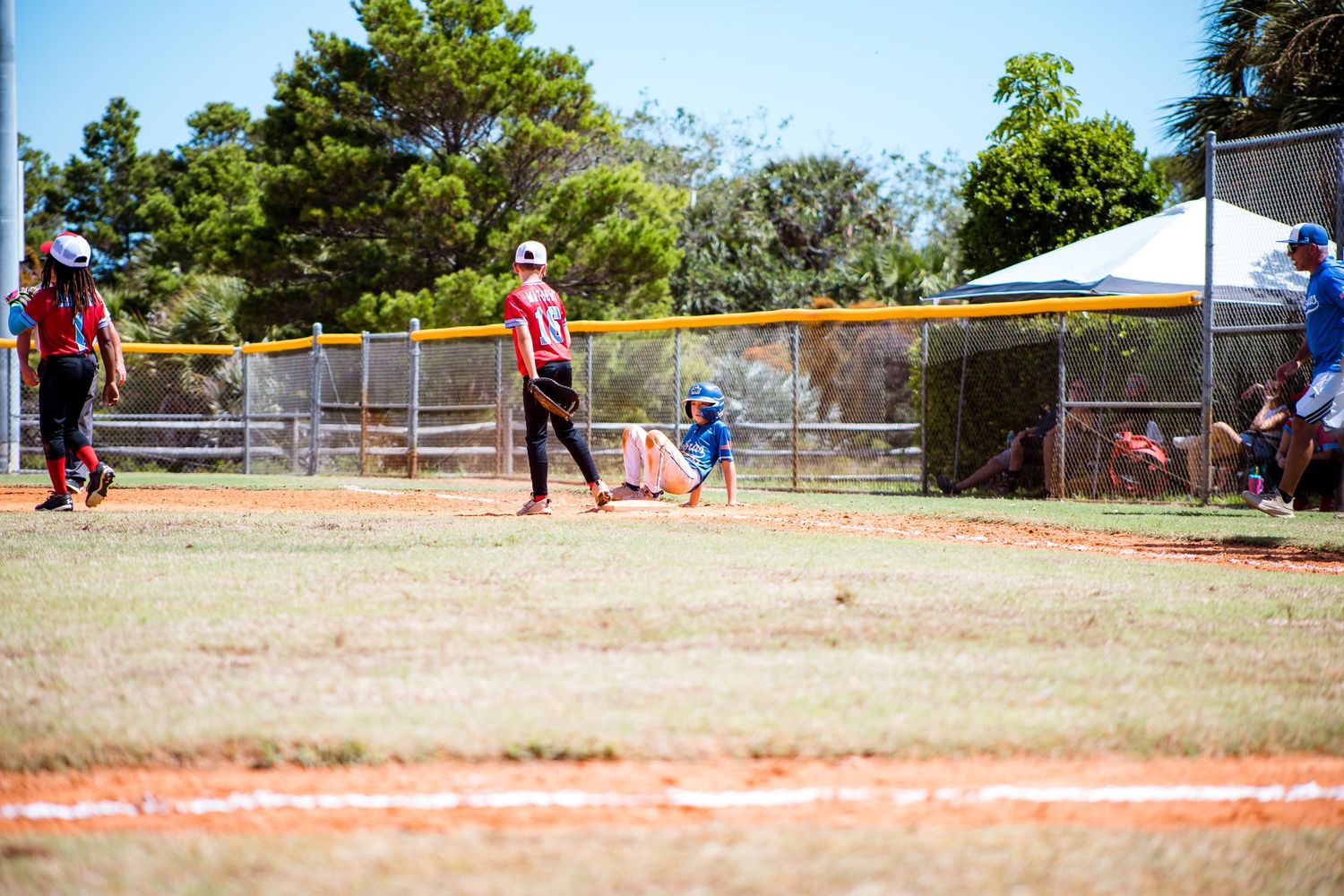 Youth baseball game in progress on sunny day with teams in red and blue uniforms.