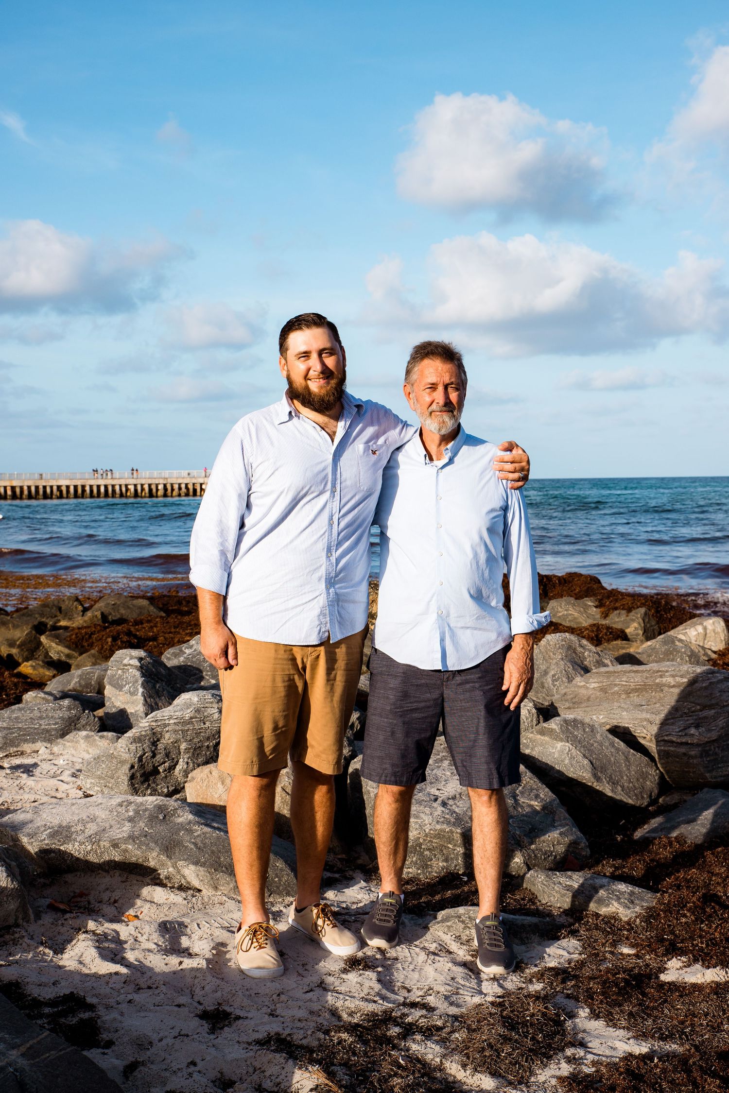 Two people stand together on rocky coastal shore during a sunny day with ocean and pier in the background.