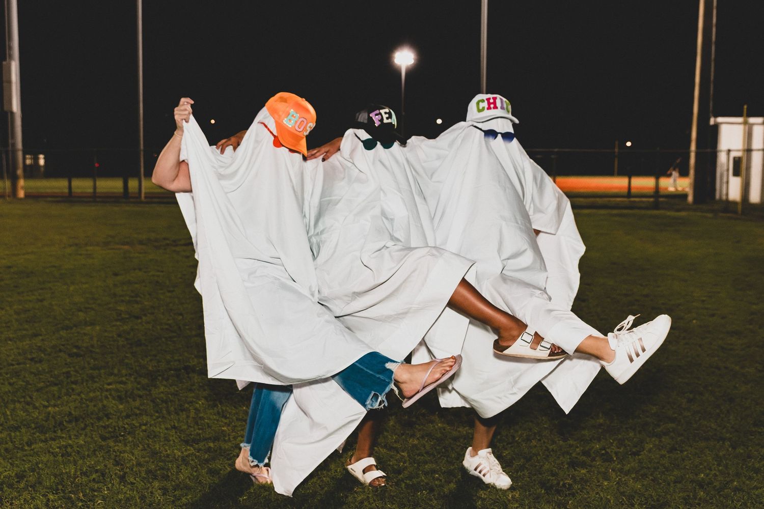 Group of people in white sheet ghost costumes gathered on a dark field with stadium lights in background.