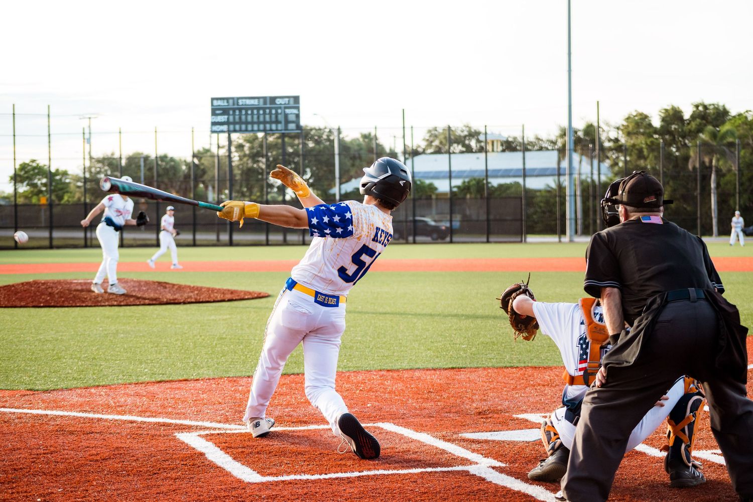 Baseball batter in white uniform takes stance at home plate during daytime game on red dirt field.