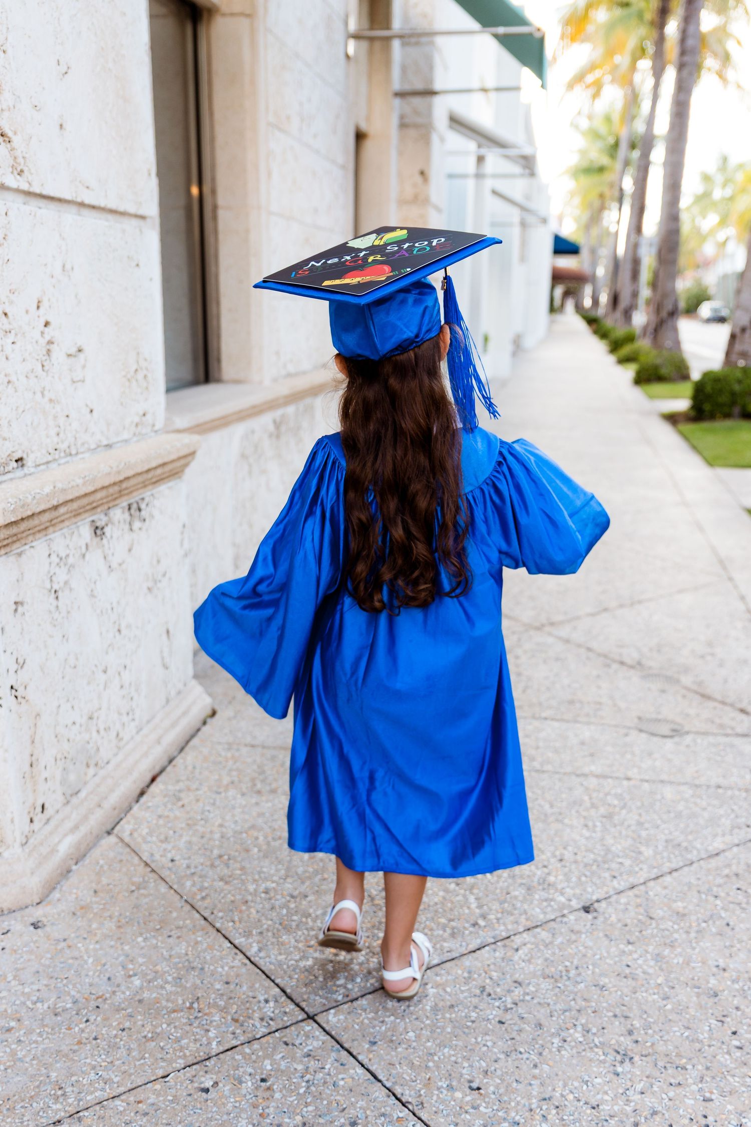 Graduate in bright blue cap and gown walks down a palm-lined sidewalk with decorated mortarboard.