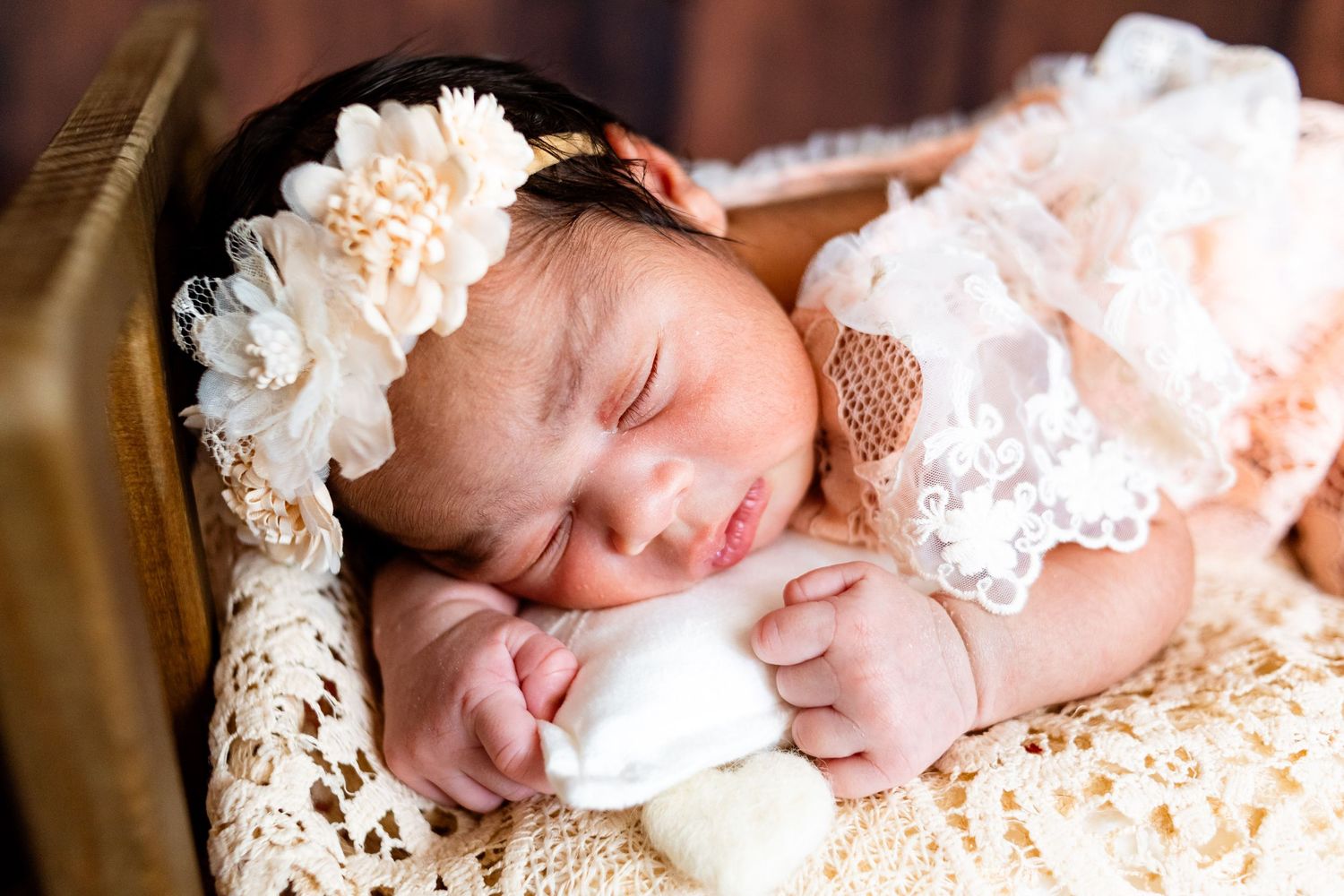 Sleeping newborn baby girl in peach lace dress and floral headband resting on crochet blanket in wooden box.