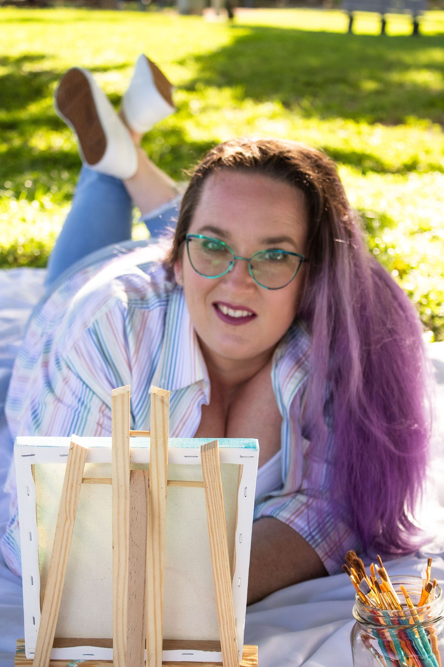Artist lying on blanket with easel and art supplies in sunny outdoor setting.