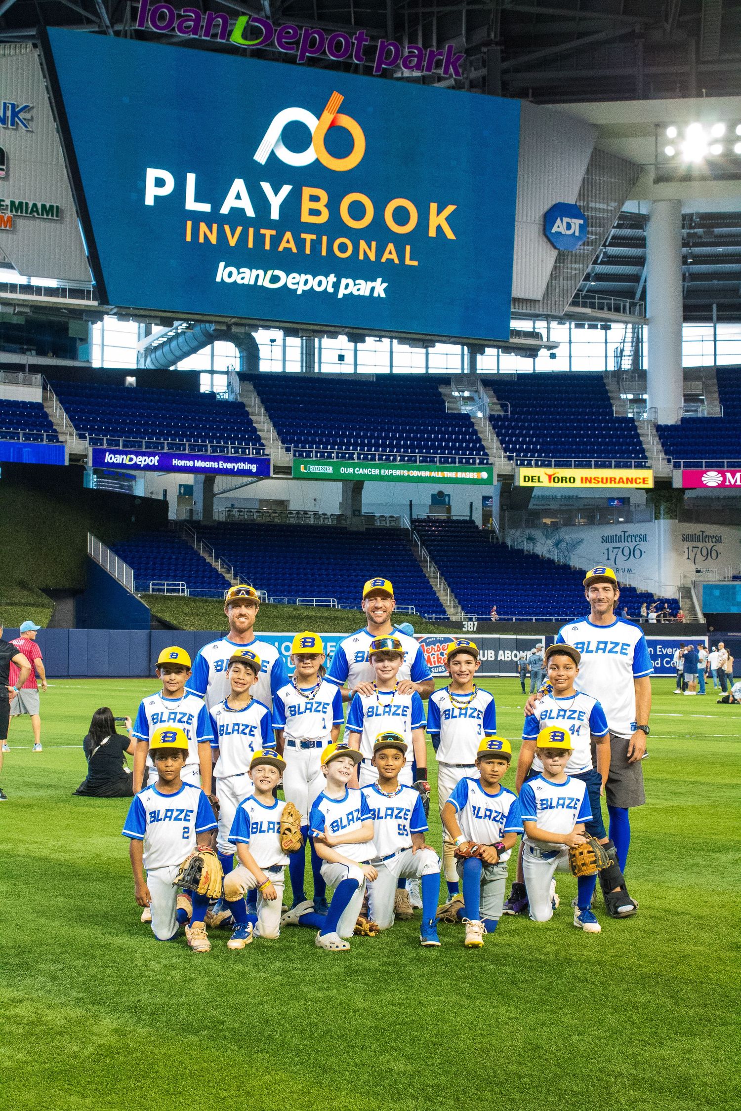 Team photo of youth baseball players in blue and white uniforms on field under Playbook Invitational sign.
