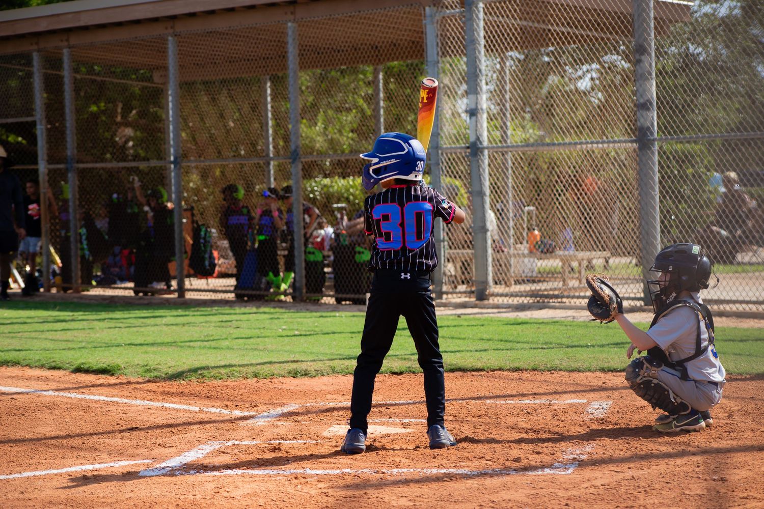 Baseball batter at plate with catcher and umpire during game under covered dugout.