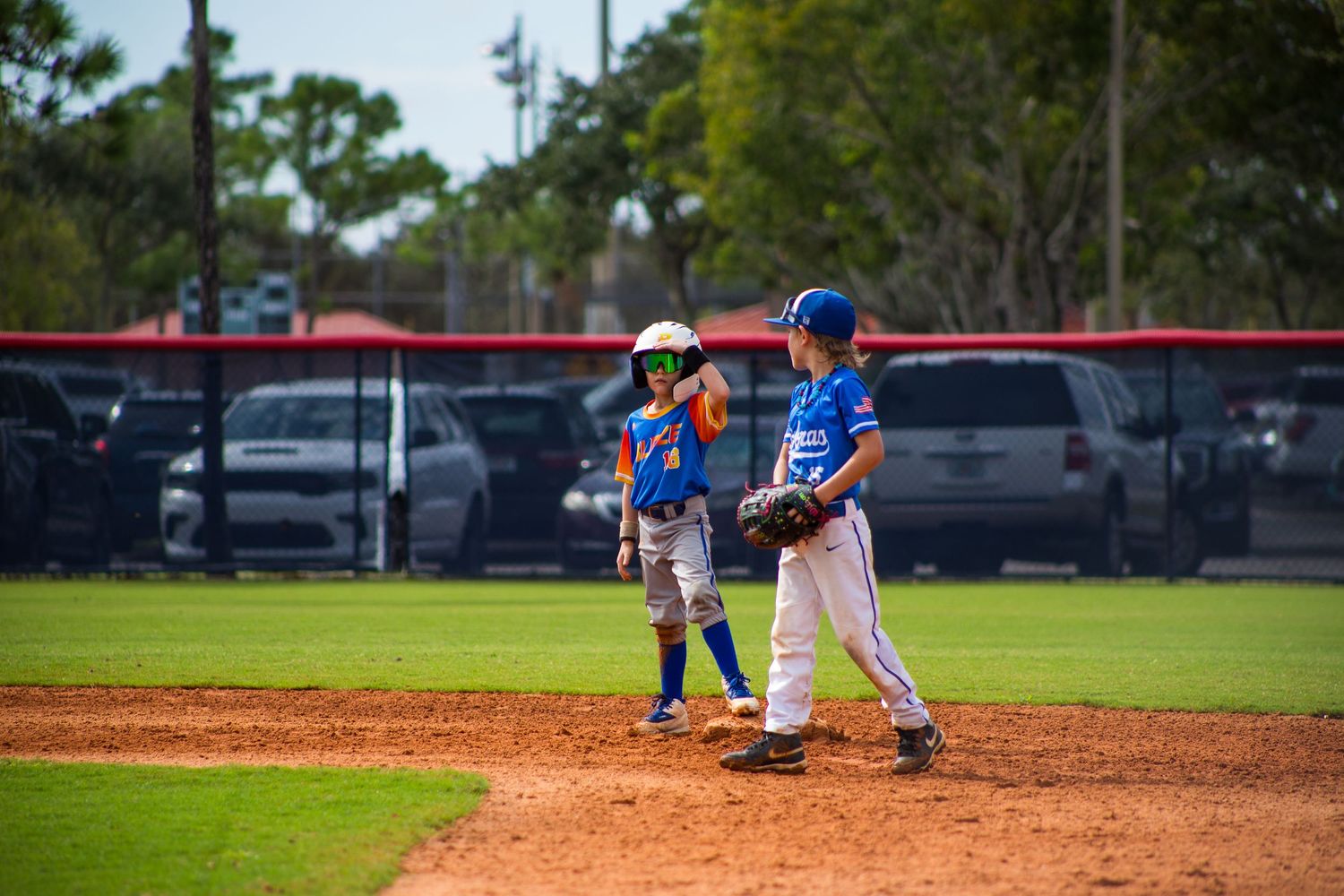Two youth baseball players in blue uniforms interact on the pitcher's mound during a game.