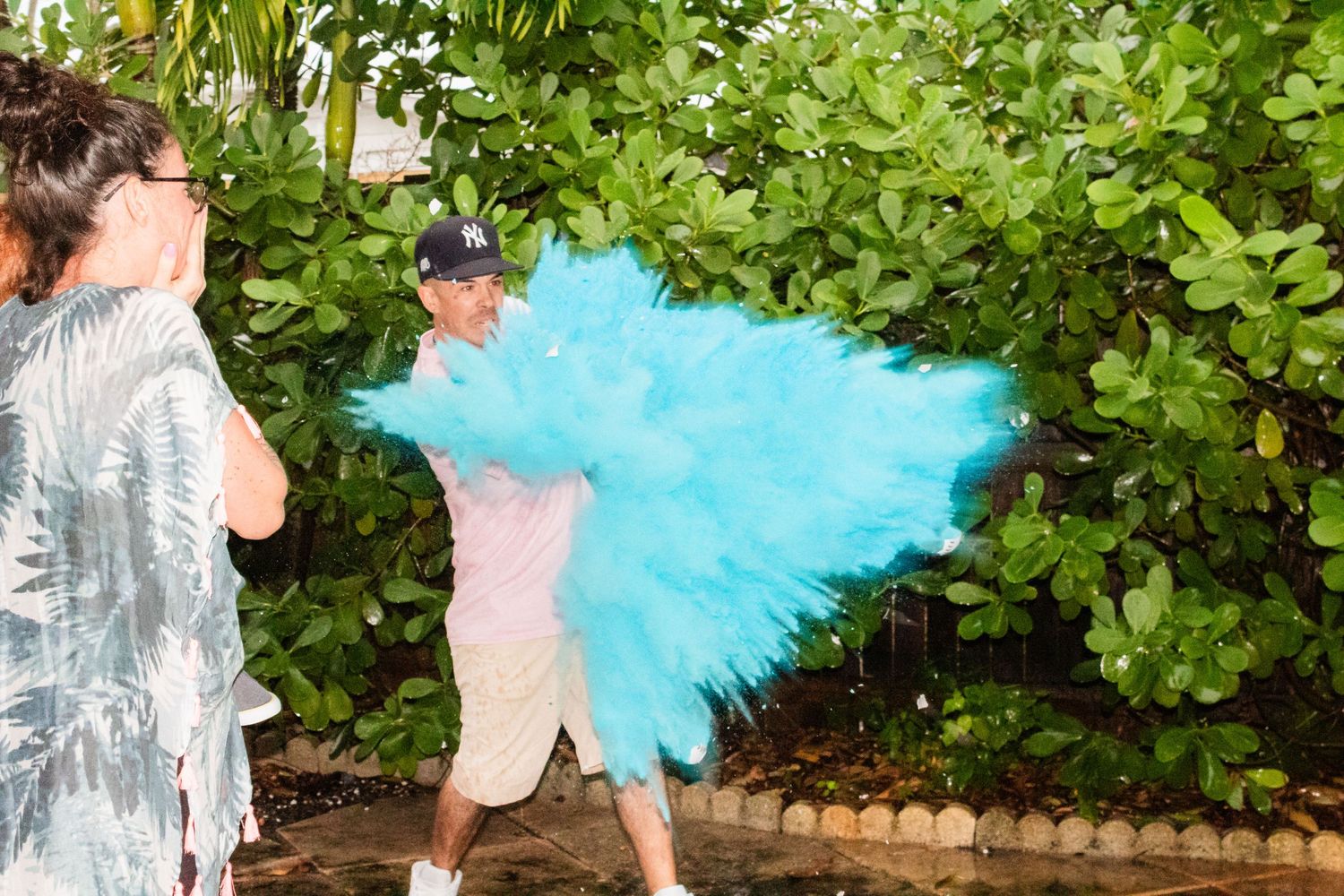 Outdoor gender reveal moment with bright blue smoke effect against green foliage.