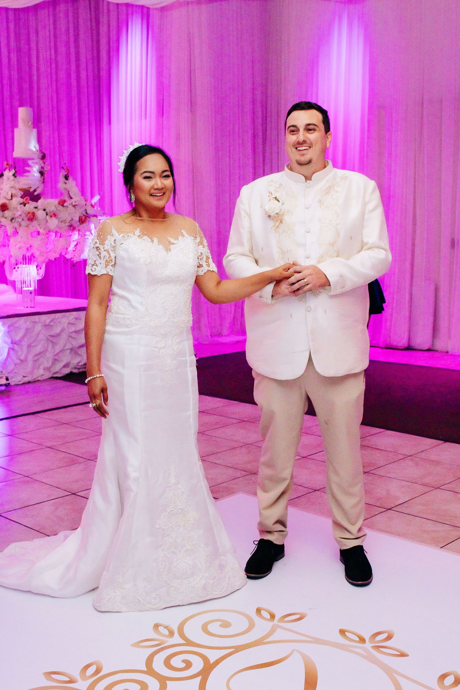 Couple stands together on decorated dance floor with pink lighting and floral arrangements.