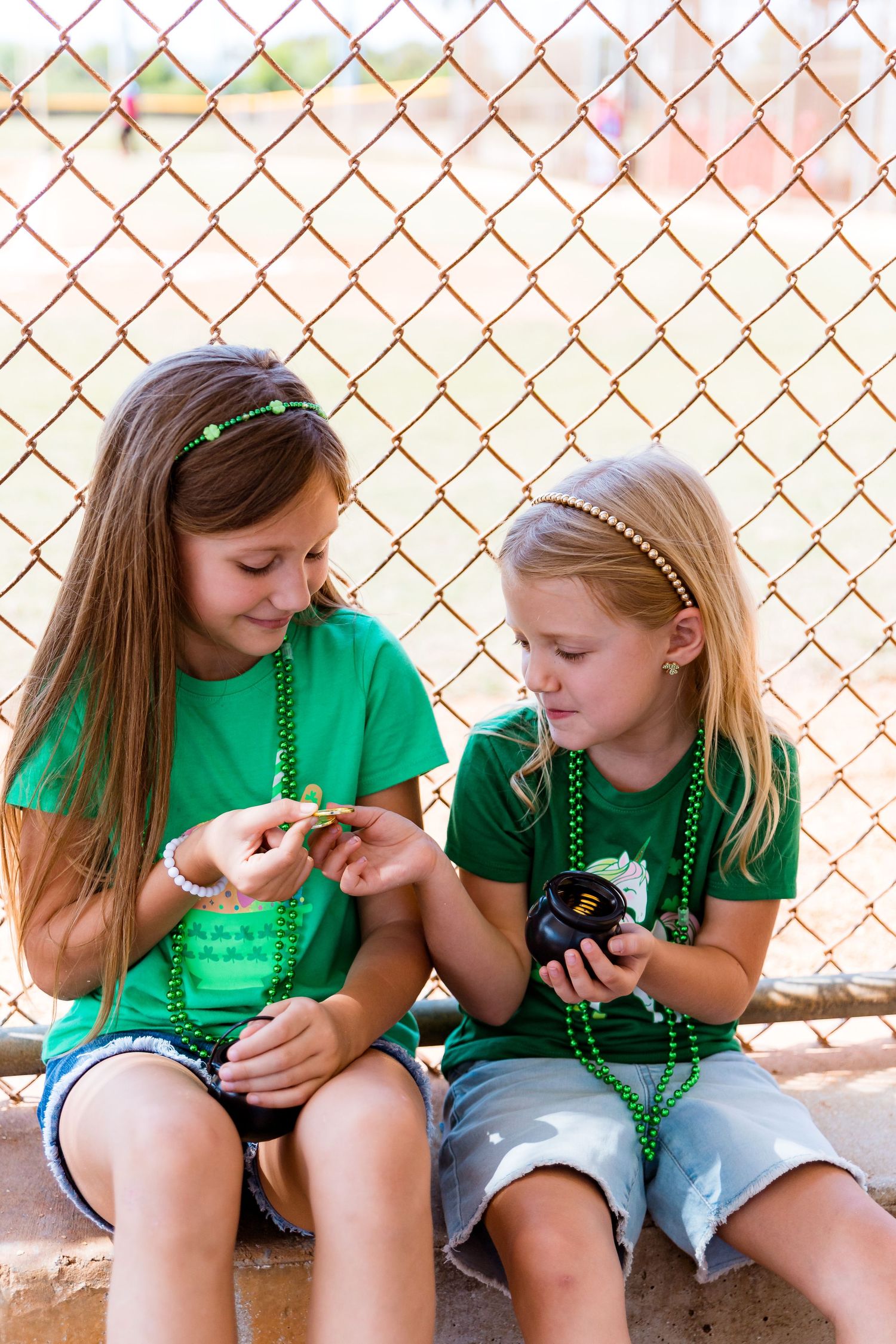 Two children in green shirts sitting with beads against chain link fence.
